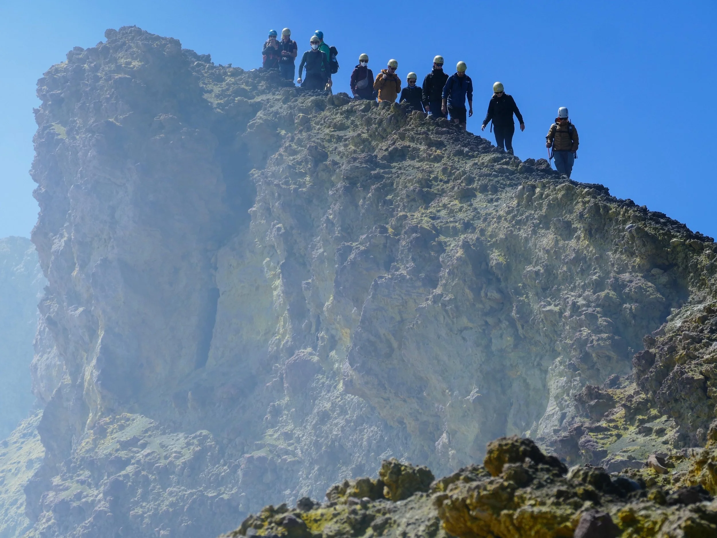 Gruppo di escursionisti con caschi che attraversano una cresta rocciosa in montagna con cielo azzurro sullo sfondo.