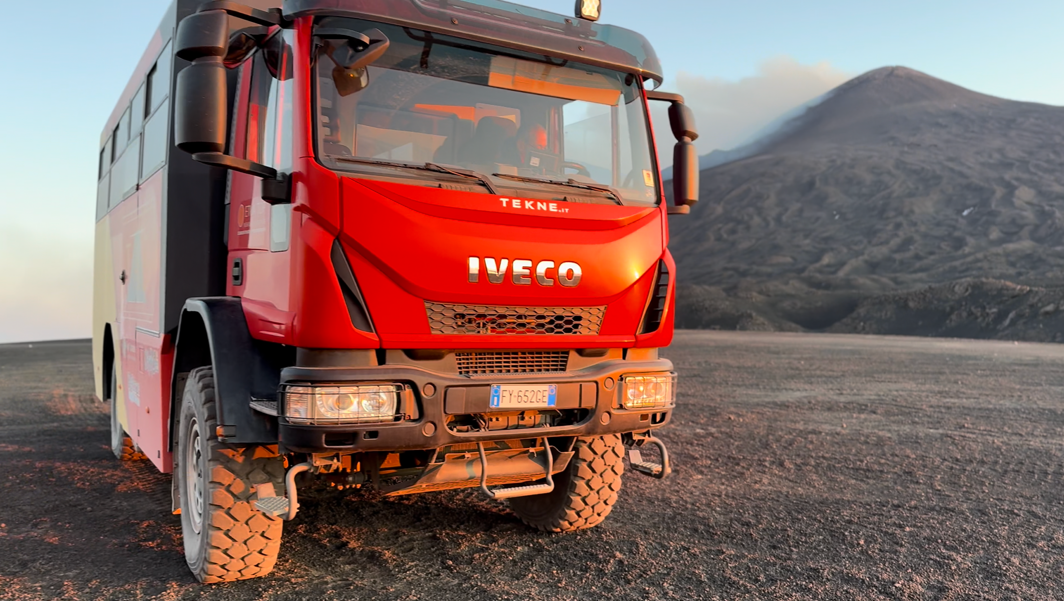Camion rosso con il logo IVECO e il sito TEKNE.IT, in un paesaggio con montagne al tramonto.