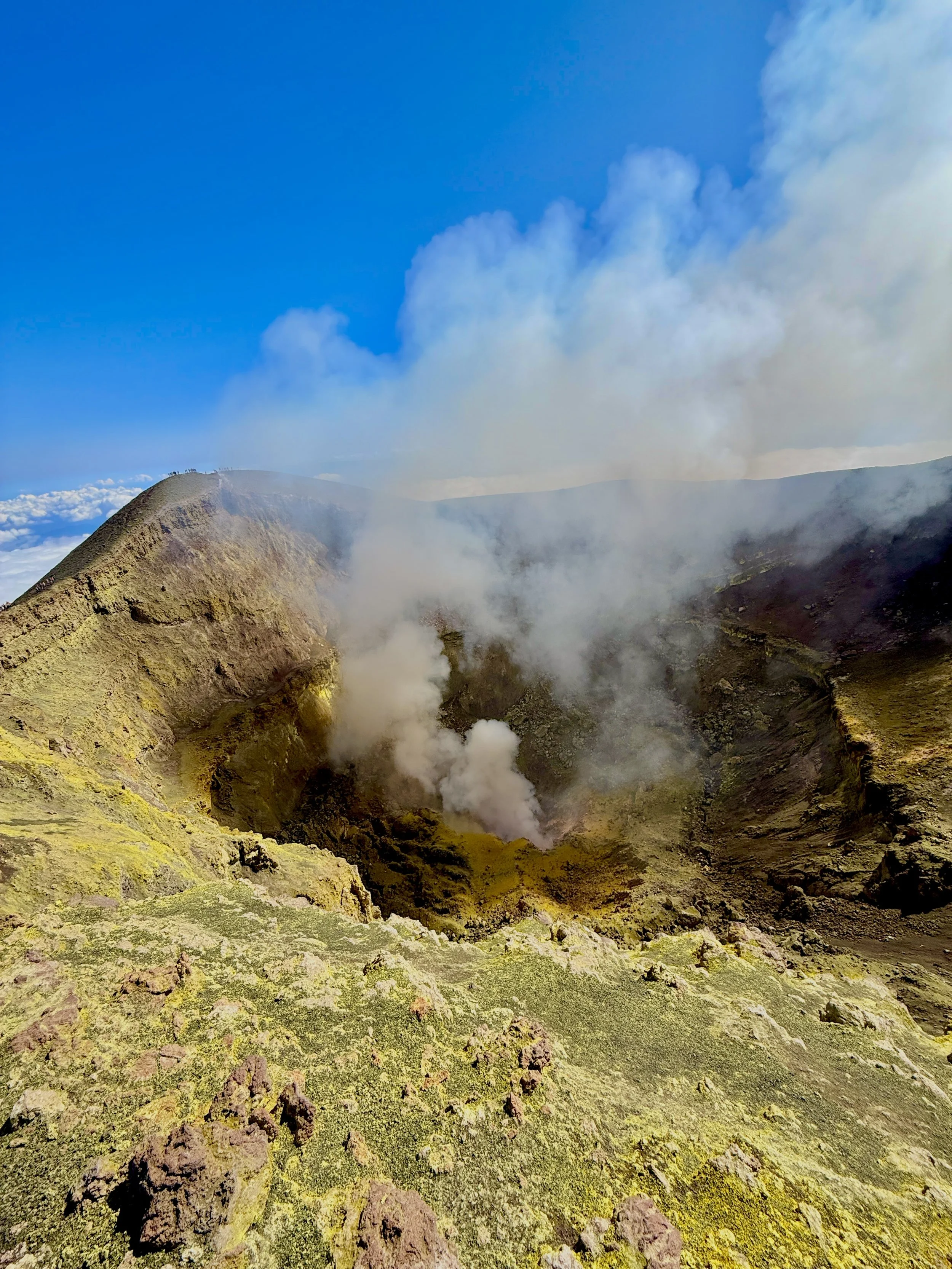 Un cratere vulcanico con fumarole che emettono vapori, su un paesaggio roccioso e ricoperto di licheni verdi, sotto un cielo blu con alcune nuvole.