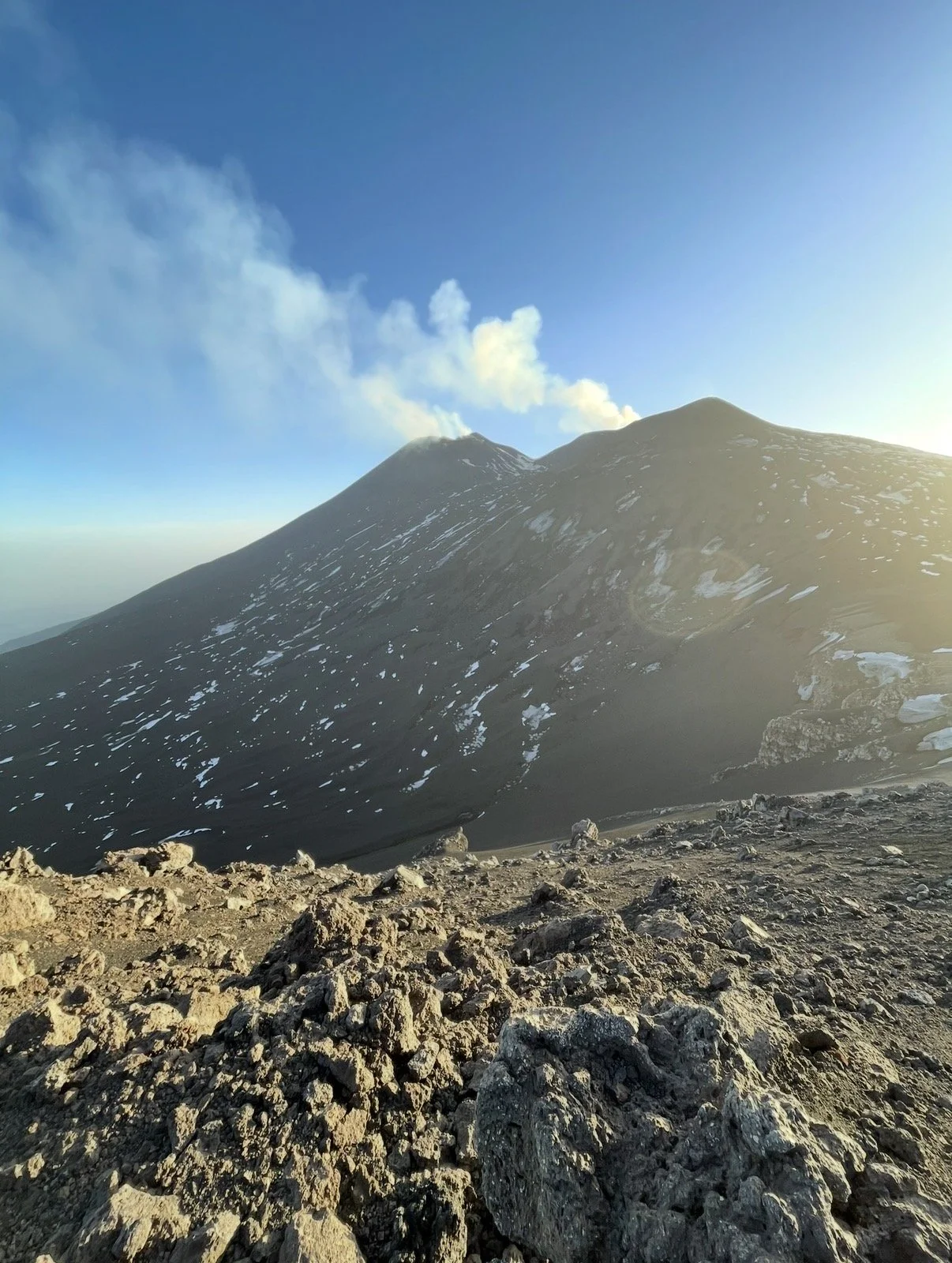 Un vulcano che emette fumaraci dalla sommità, con un paesaggio roccioso e sterrato e un cielo blu con alcune nuvole.