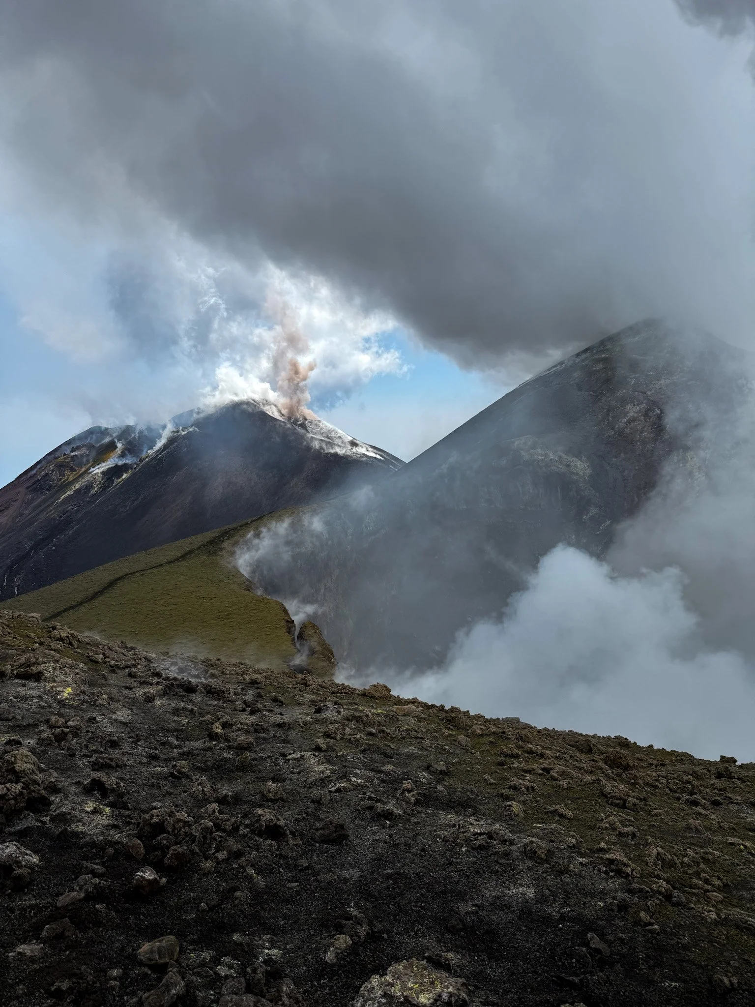 Eruzione di un vulcano attivo con colonna di fumo e lava, circondato da un paesaggio roccioso e nuvole di fumo.