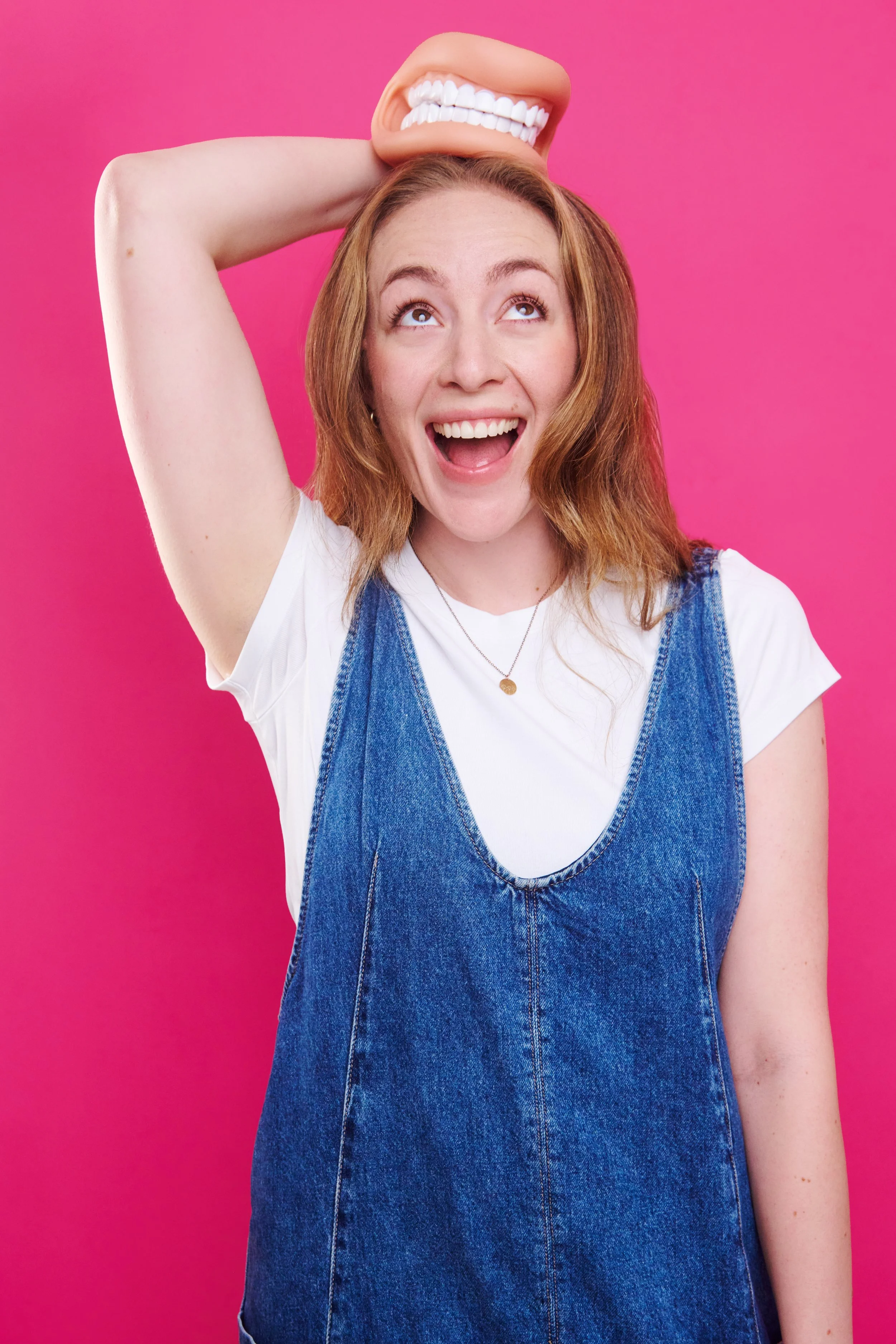 A woman with light skin, wavy blond hair, wearing a white t-shirt and denim jumper, holding a large plastic mouth with a set of teeth on her head against a pink background, smiling and looking up.