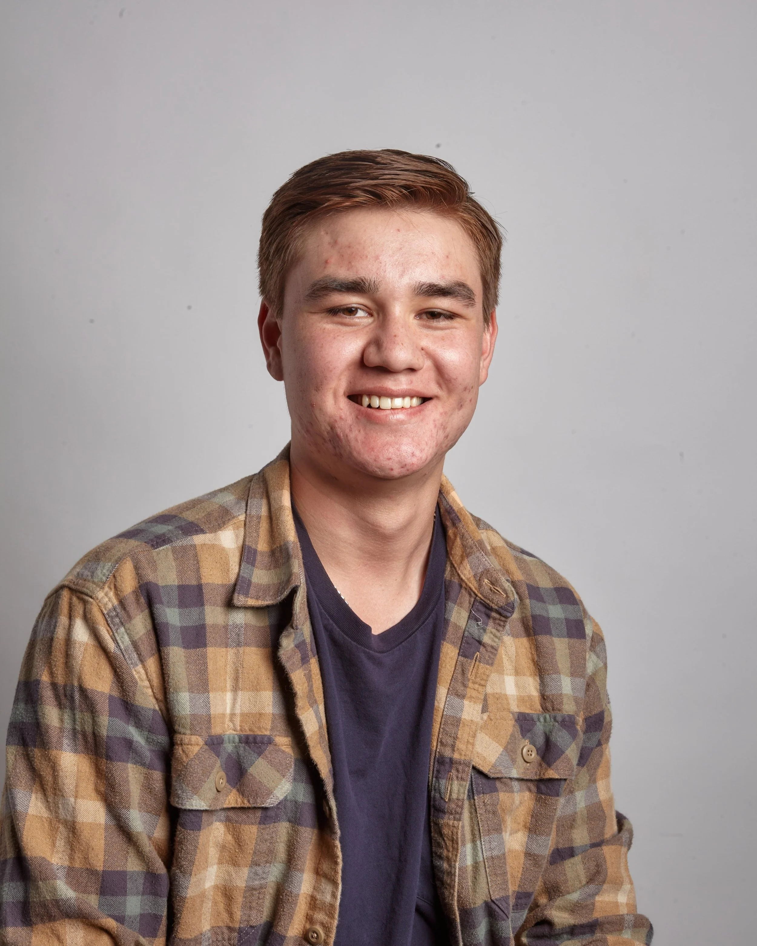 A young man with brown hair, smiling, wearing a checkered shirt over a navy T-shirt, posed against a plain gray background.
