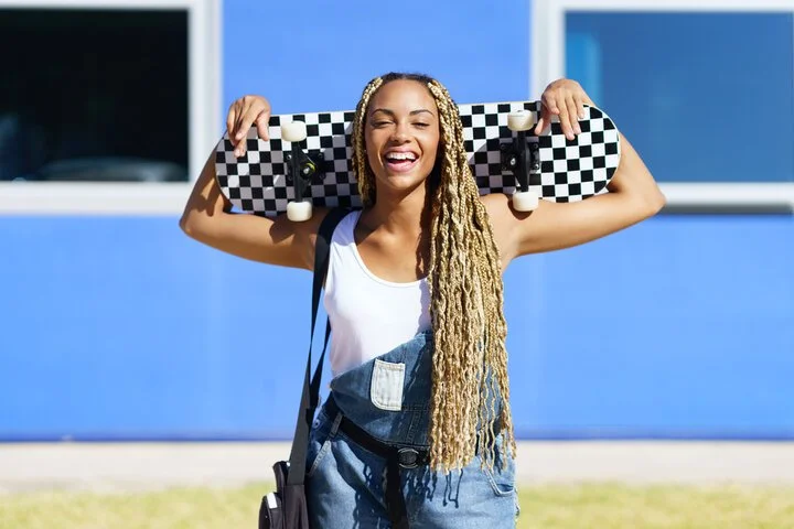 Young woman with long blonde dreadlocks smiling and holding a skateboard on her shoulders outside on a sunny day.