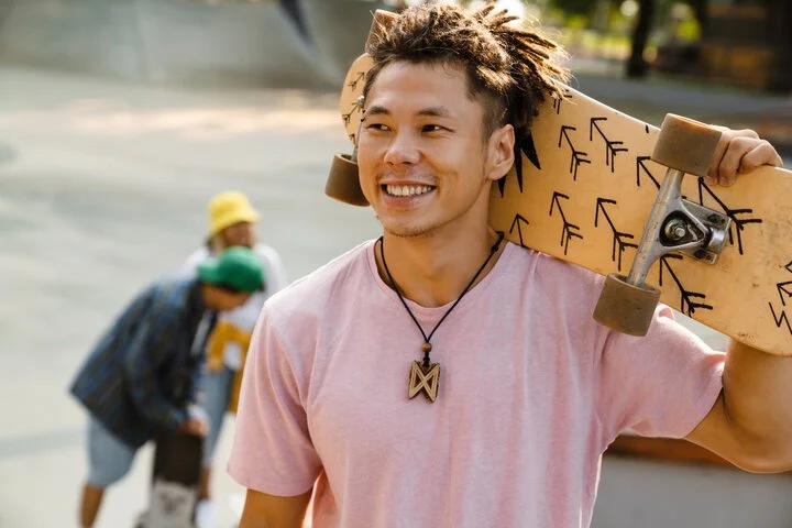 A young man with dreadlocks smiling while holding a skateboard on his shoulder in an outdoor setting with a blurred background of other people.