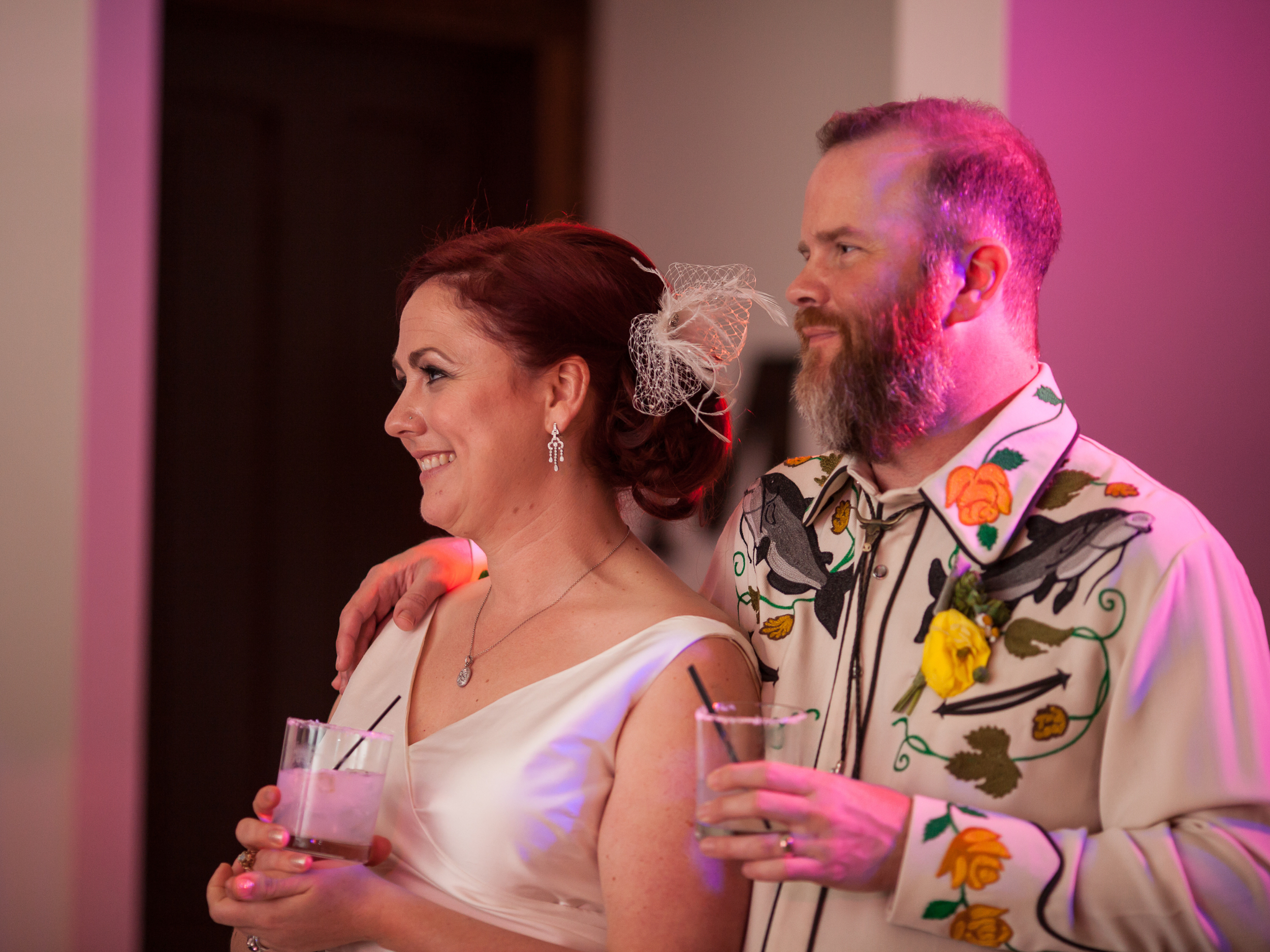 A woman with red hair and earrings smiling, holding a drink, and a man with a beard, wearing a colorful embroidered shirt, also holding a drink, at a celebration or party.