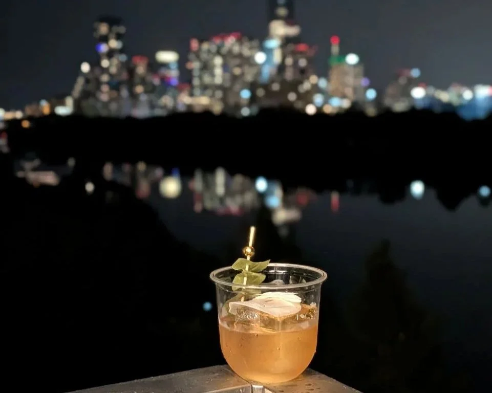 A cocktail with ice in a clear plastic cup, garnished with a lime wedge and a fancy stir stick, set against a blurry city skyline at night.