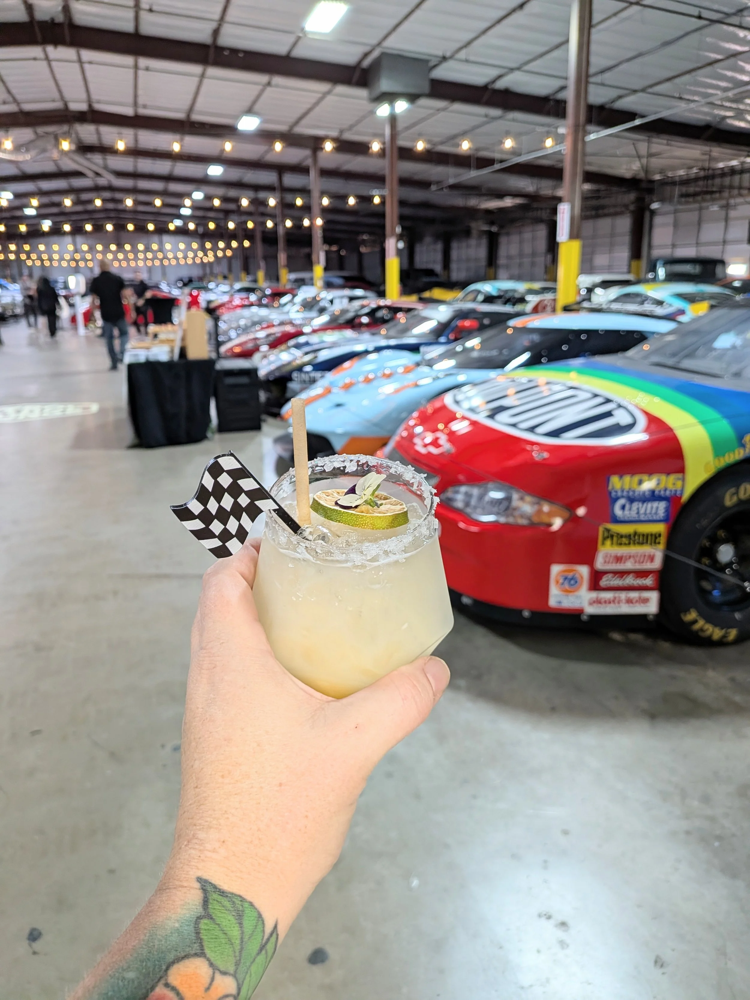 A hand with a colorful tattoo holding a cocktail with lime and lemon slices and a checkered flag garnish in front of a collection of race cars inside a large indoor car storage or display facility.