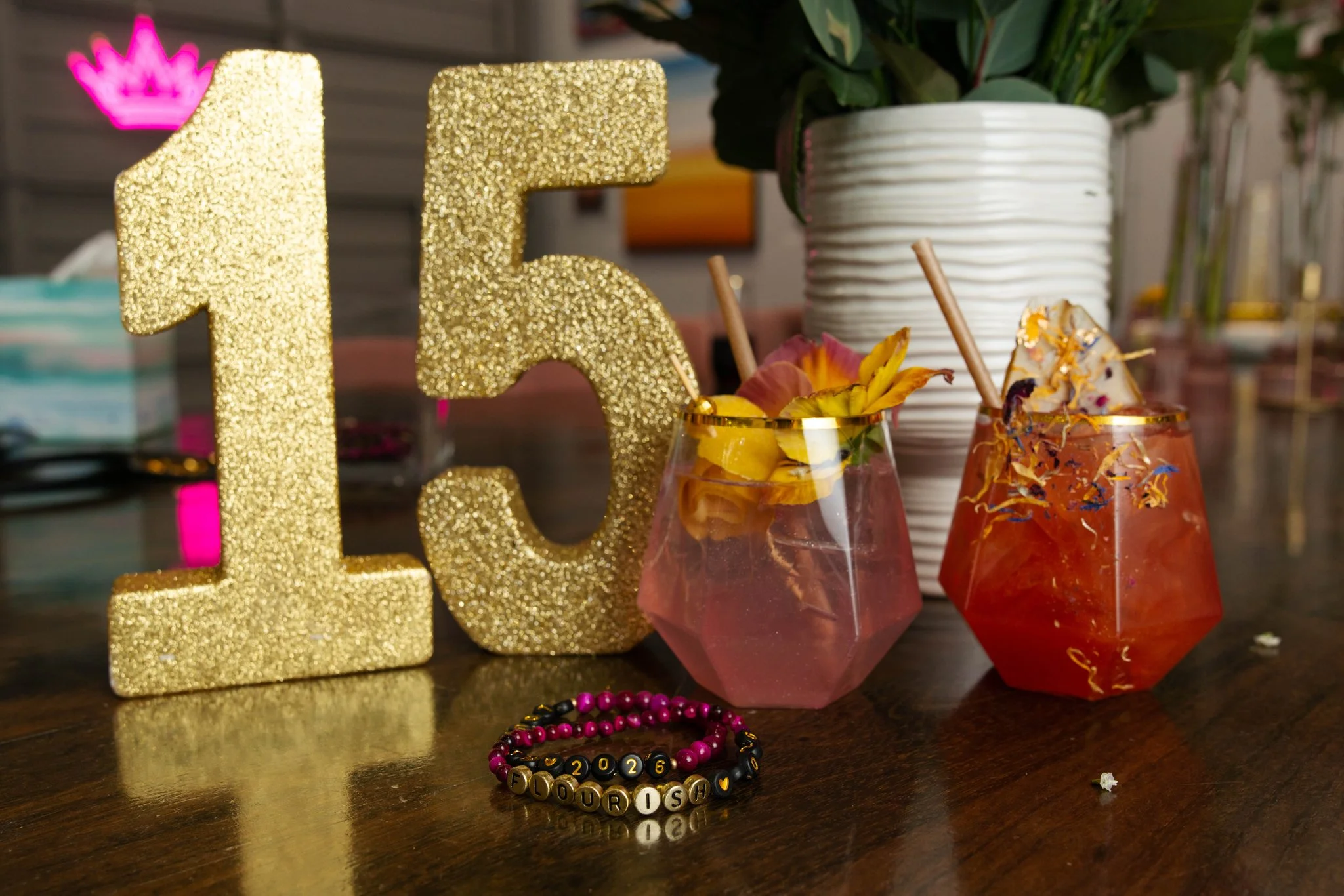 Gold glitter number '15' candle, two cocktails with flower garnishes and colorful decorations, a white vase with green leaves, a beaded bracelet with '2026' and 'FLOURISH' spelled on it, on a dark wooden table.