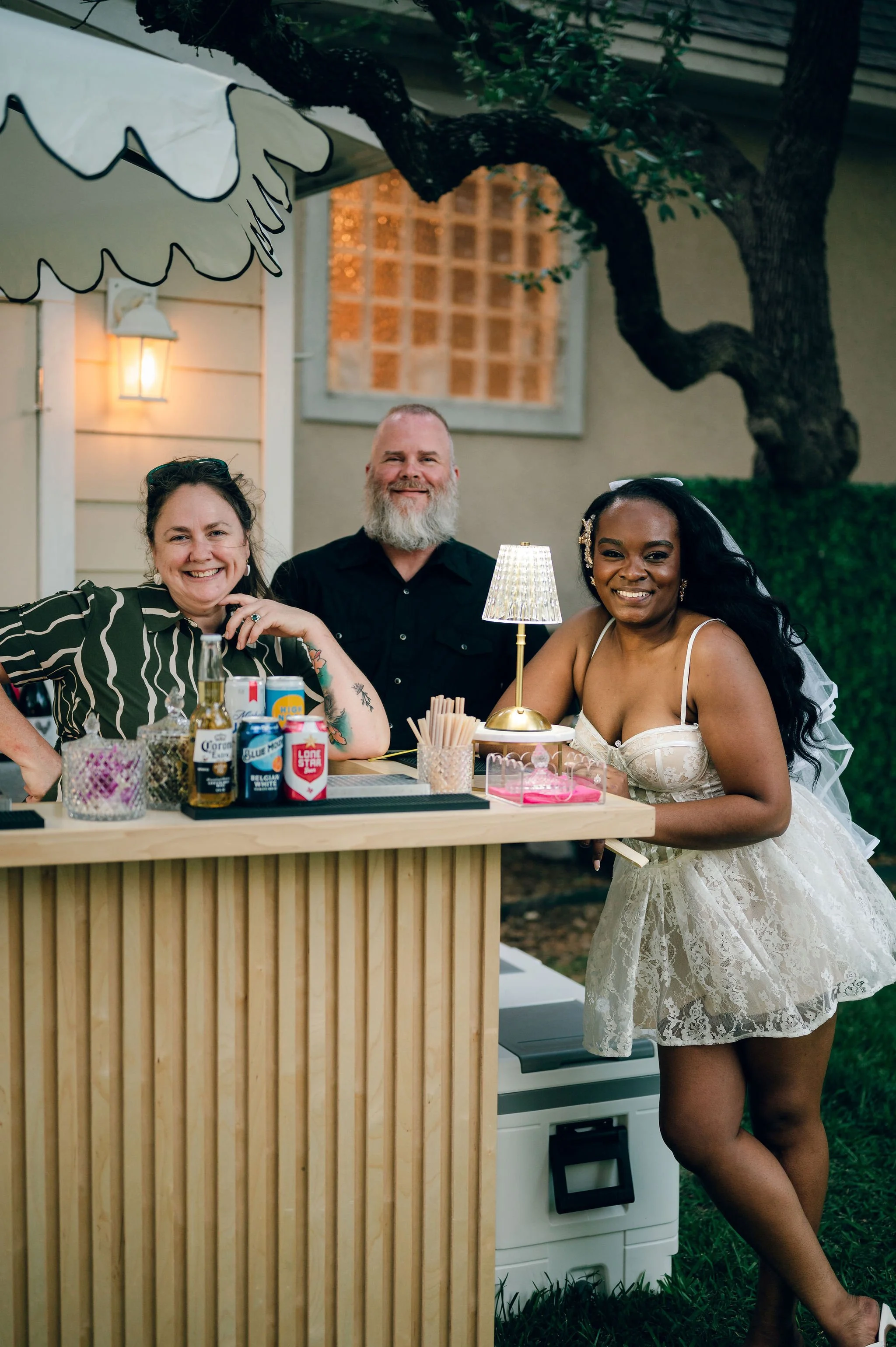 A woman in a white lace dress and a veil standing next to a bar at an outdoor gathering, smiling at the camera. A man and another woman sit behind the bar, smiling, with drinks and bottles on the bar. The setting is outside with a tree, a building, a