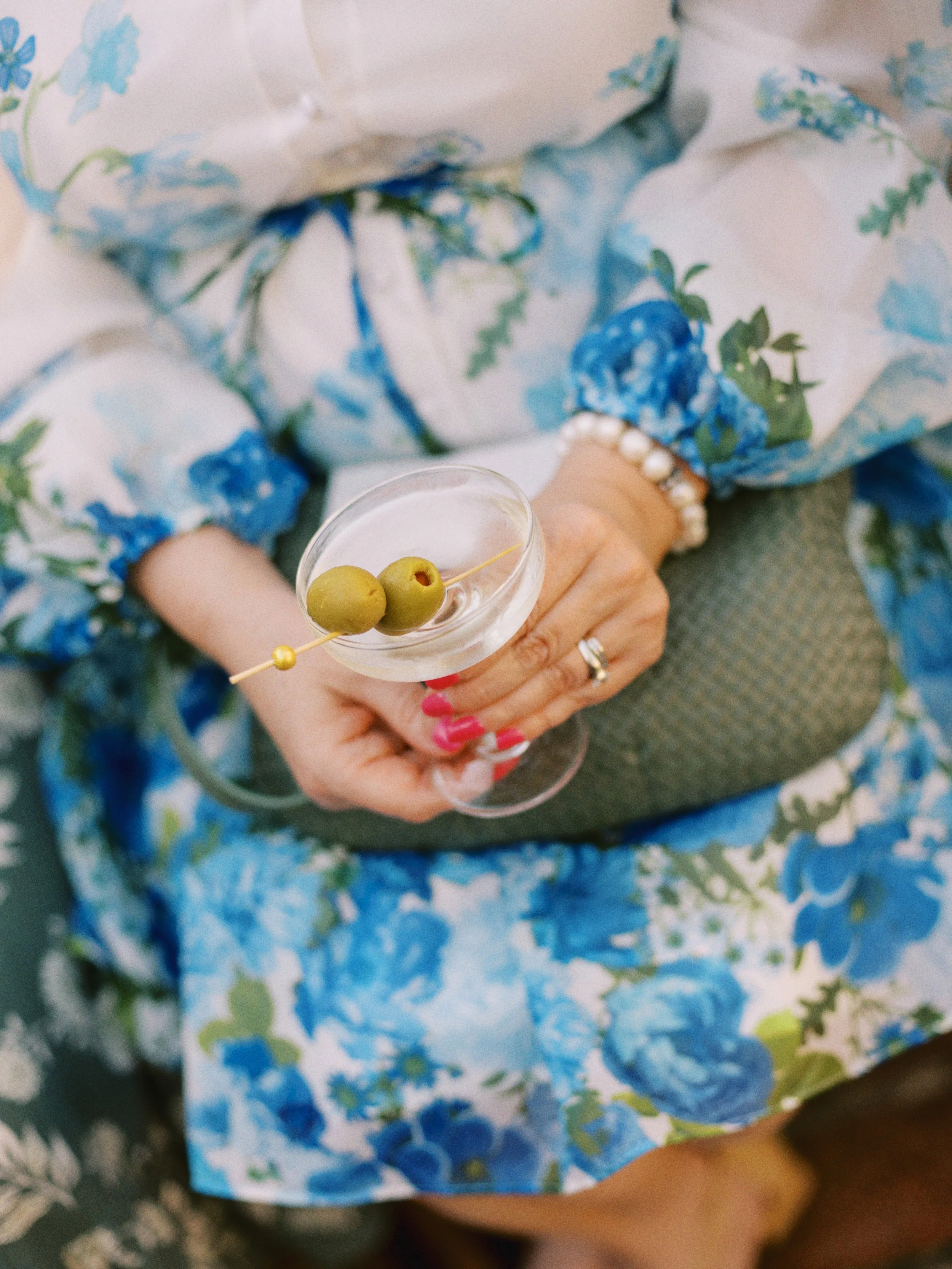 Person holding a martini glass with green olives and toothpick, wearing a blue floral dress with pearl bracelet.
