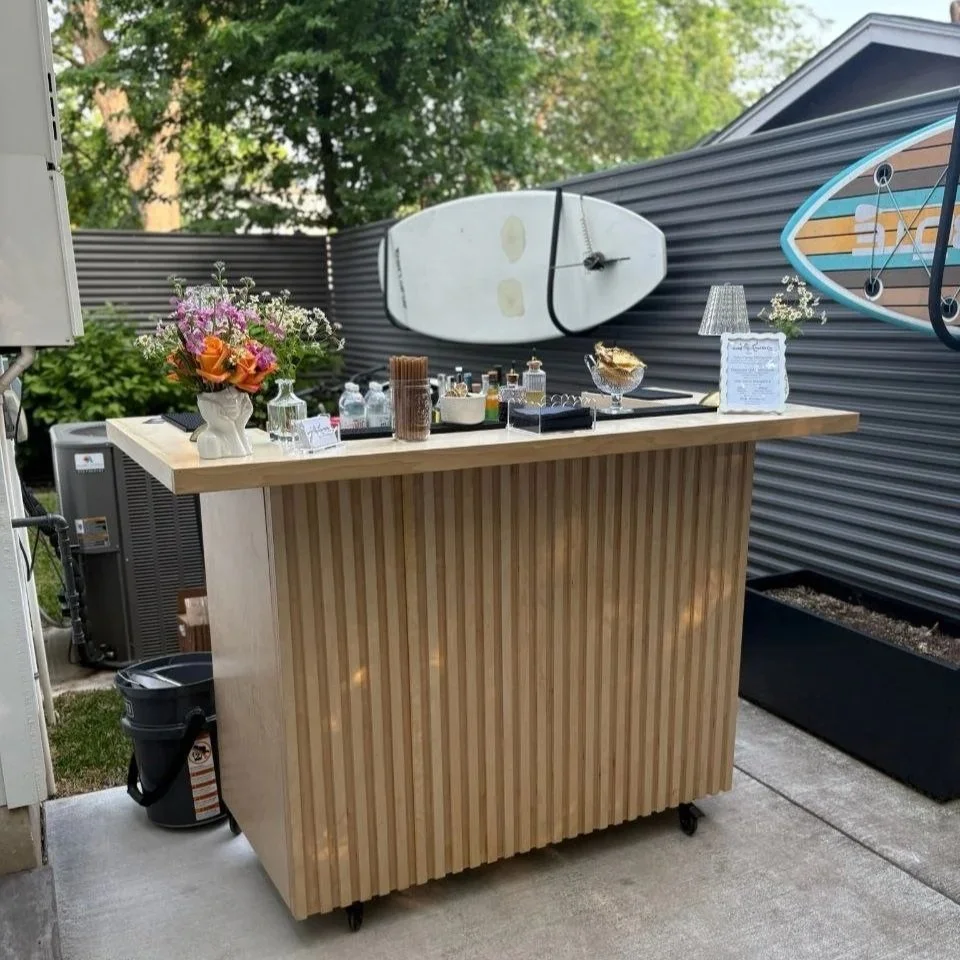 Social Butterfly ATX mobile bar cart set up at an Austin backyard event, styled with fresh florals, a printed cocktail menu, and bar tools against a surfboard backdrop