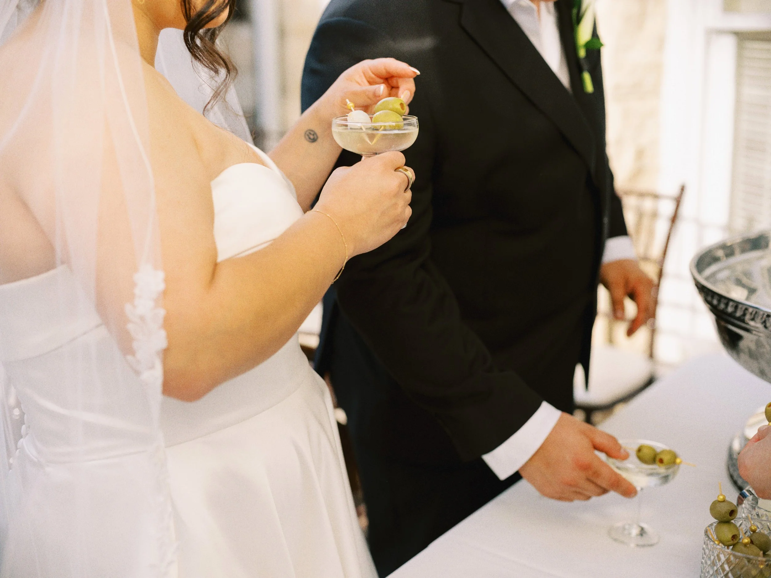 Close-up of a bride in a white dress holding a cocktail glass with grapes, standing next to a groom in a black suit at a reception table.