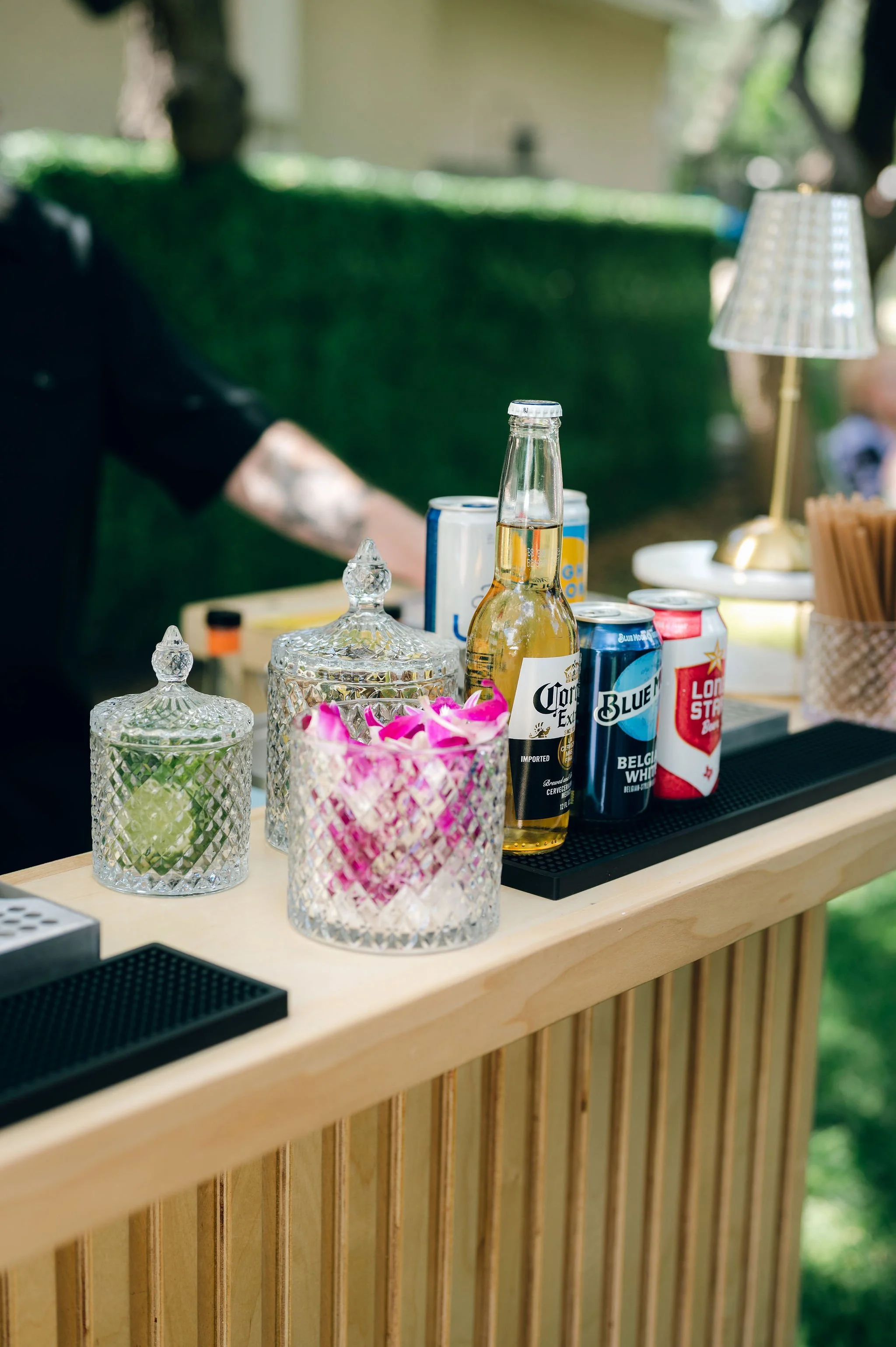 A bar setup with various beverages including beer and soda cans, glass jars with lids, a small decorative vase with pink and white flowers, and a table lamp, all on a wooden bar counter with vertical slats, in an outdoor setting with blurred greenery
