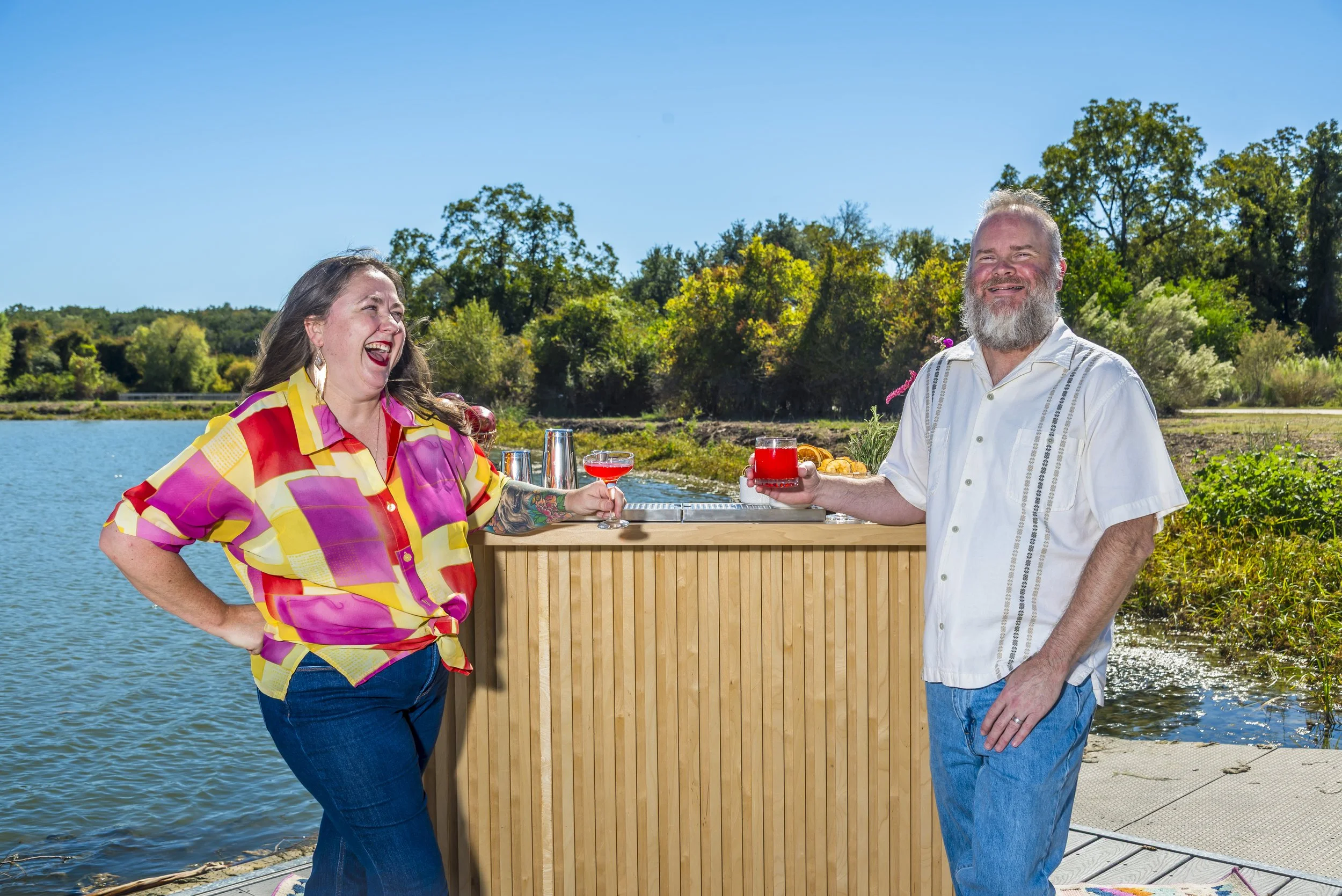 Two people, a woman and a man, standing by a wooden outdoor bar near a body of water with trees in the background, smiling and holding colorful drinks on a sunny day.