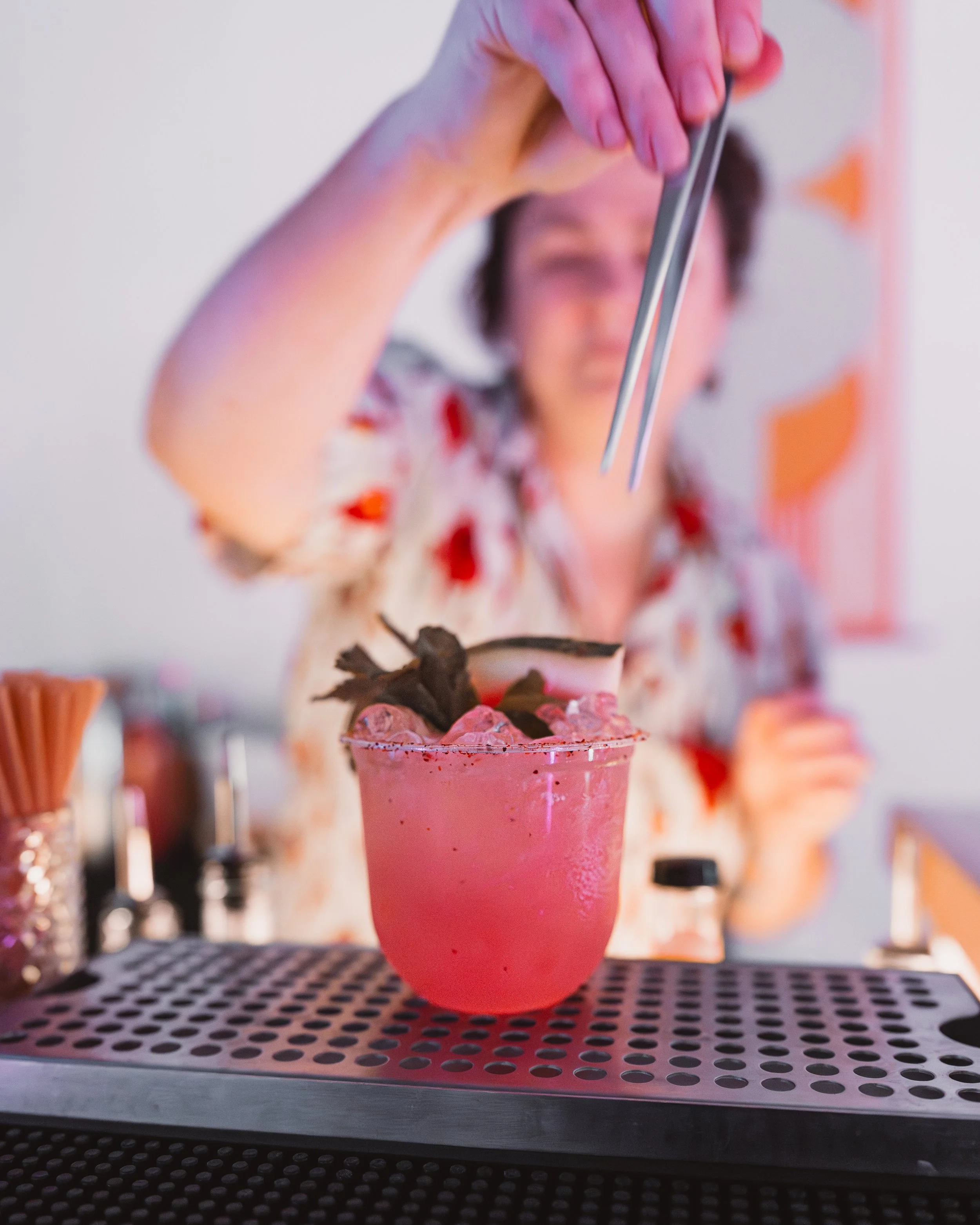 Close-up of a person pouring a pink creamy beverage into a glass with a garnish of chocolate shavings and berries, on a metal tray at a bar or cafe.