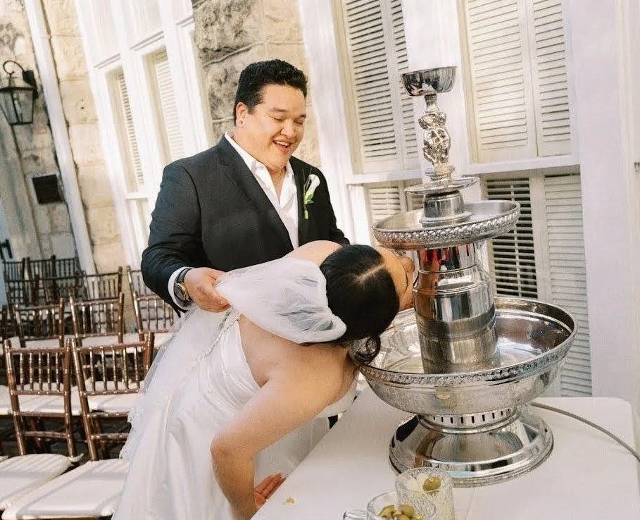 A man and a woman sharing a water fountain at a wedding reception, with the woman drinking from the fountain and the man smiling