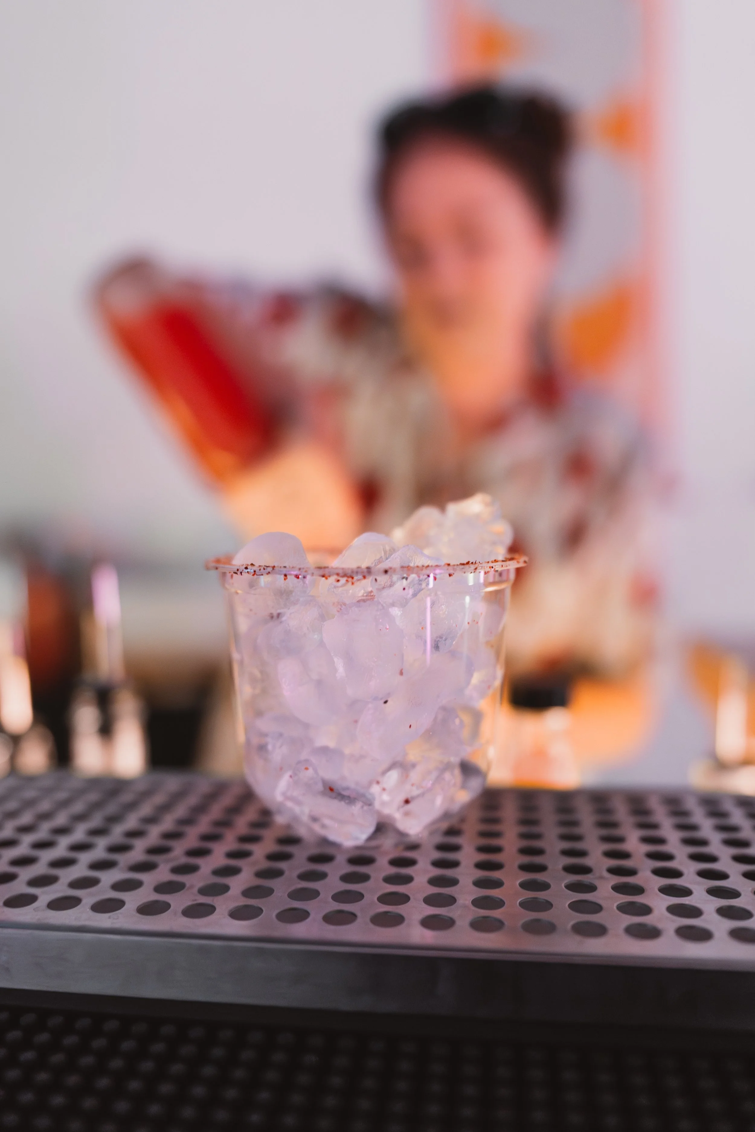 A glass filled with ice cubes on a perforated bar mat in a bar setting, with a blurred background of a bartender and bottles.