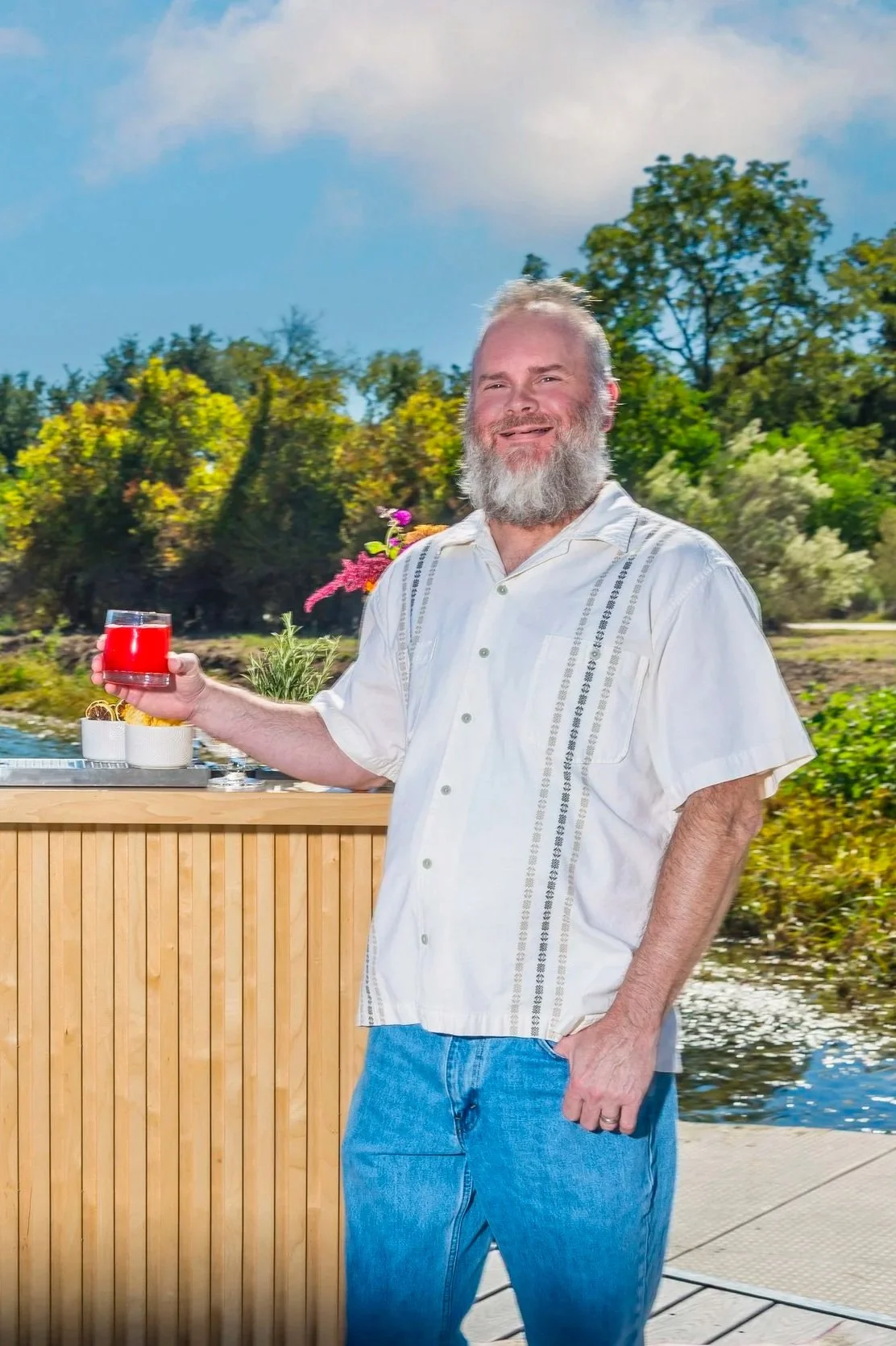 A man with a beard in a white short-sleeved shirt and blue jeans holding a red drink while standing outdoors on a sunny day near a body of water with trees in the background.