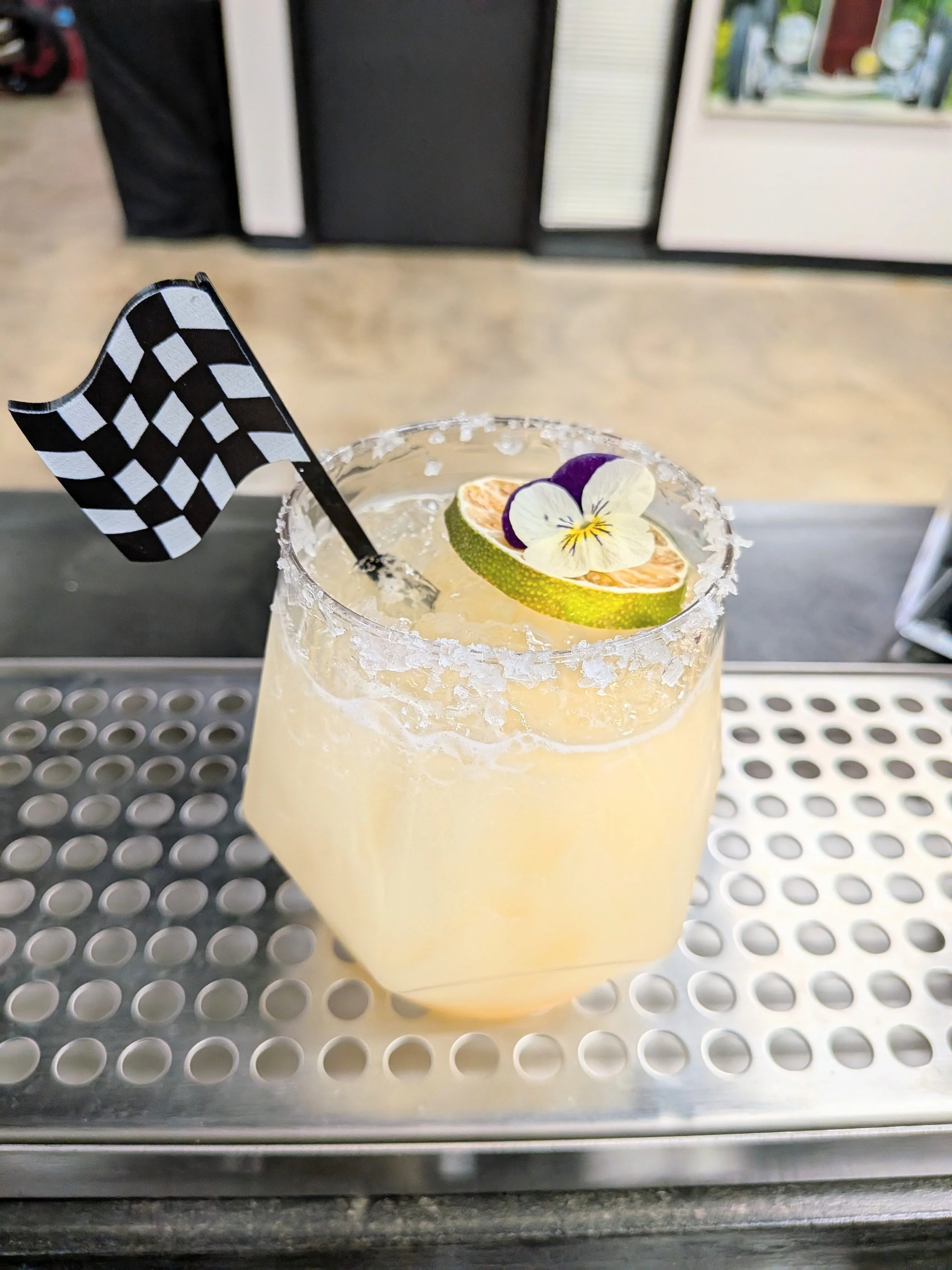 A pale yellow cocktail in a rounded glass with a salted rim, garnished with a slice of lime, a purple flower, an edible flower, and a small checkered flag, placed on a metal bar surface.