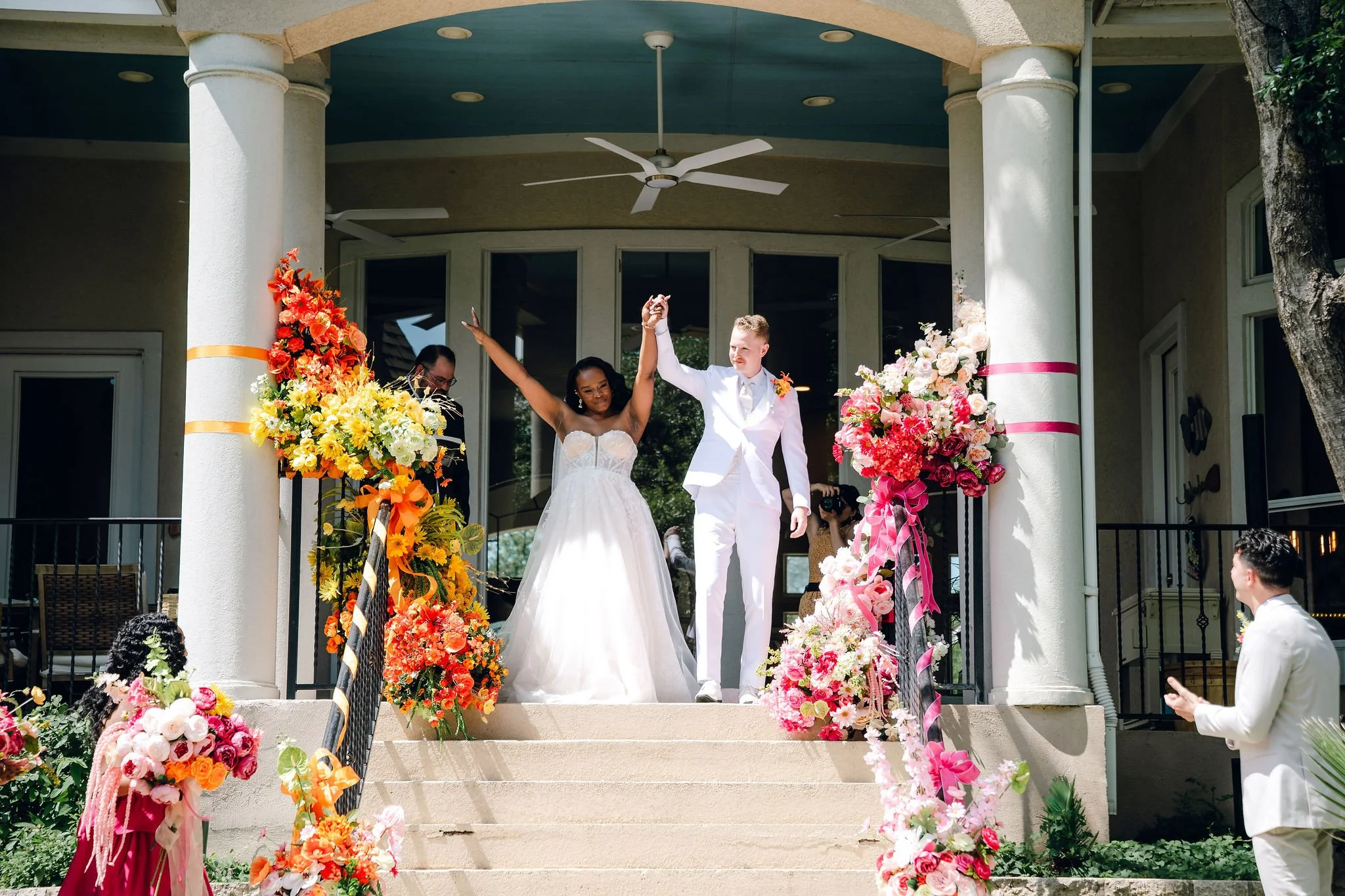 A newlywed couple celebrating on a decorated outdoor porch with floral arrangements. The bride is in a white wedding dress holding her arms up, and the groom in a white suit is holding her hand. There are vibrant flowers on either side of the porch and people gathered around, part of a wedding celebration.