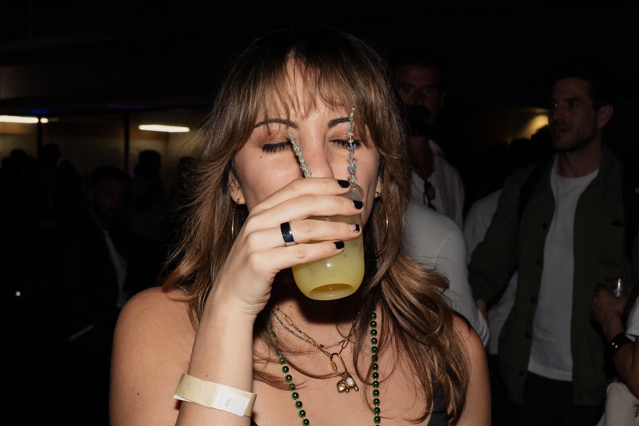 A woman with light brown hair and black nails is drinking a light yellow beverage through a straw at a party or event, with people in the background.
