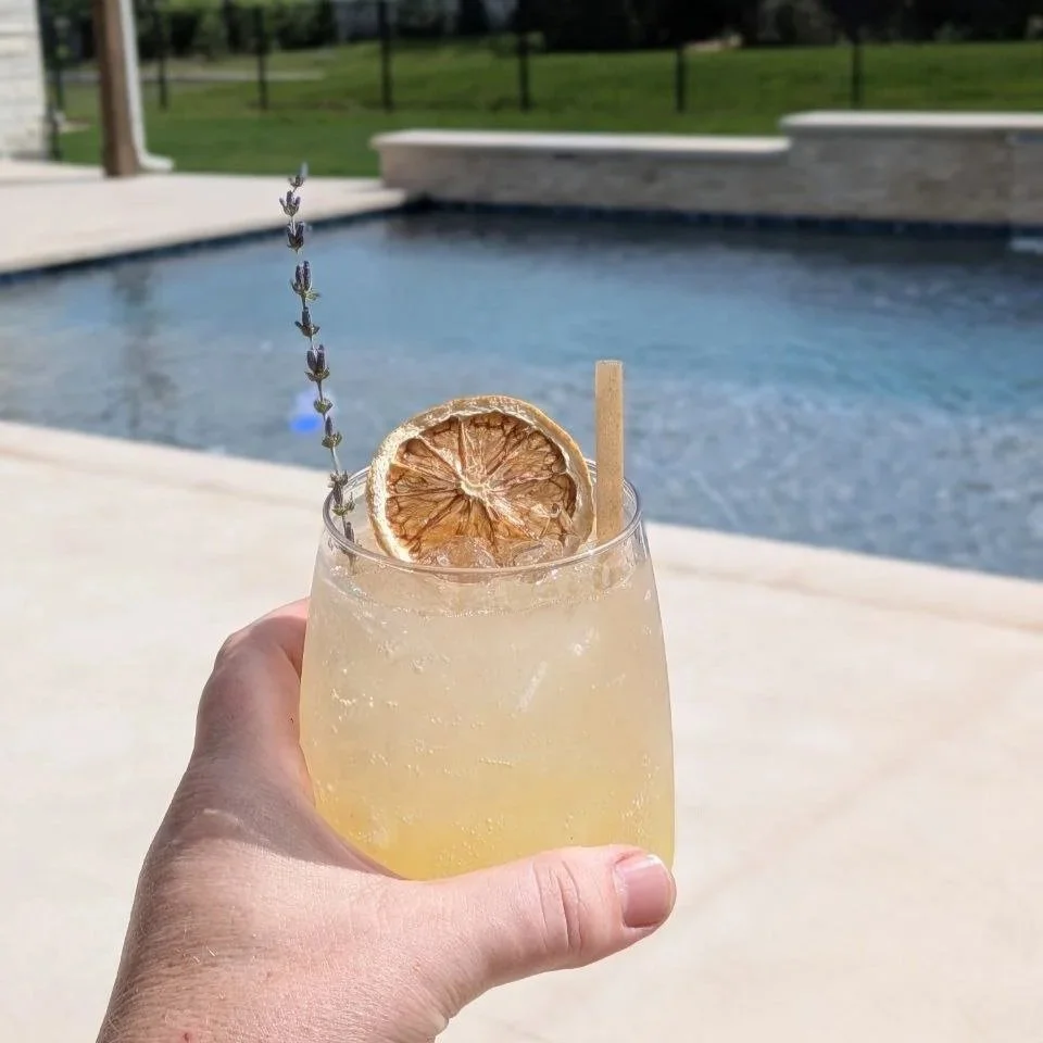 Social Butterfly ATX Lavender Oasis cocktail held poolside at an Austin backyard event, garnished with a dried citrus wheel, fresh lavender sprig, and a paper straw