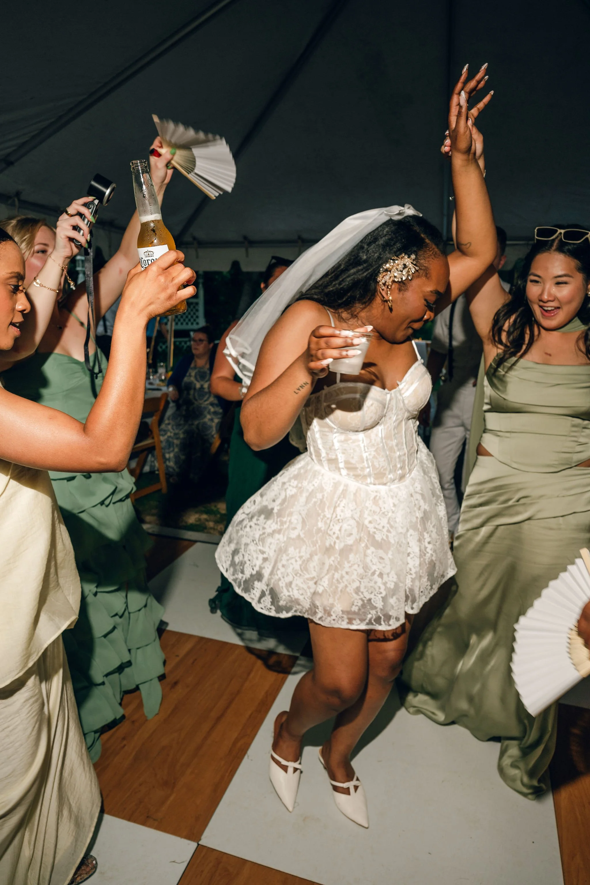 Women dancing at a celebration, with one woman in a white lace dress and veil holding a drink, surrounded by other women in green dresses, some holding fans and a beer bottle.
