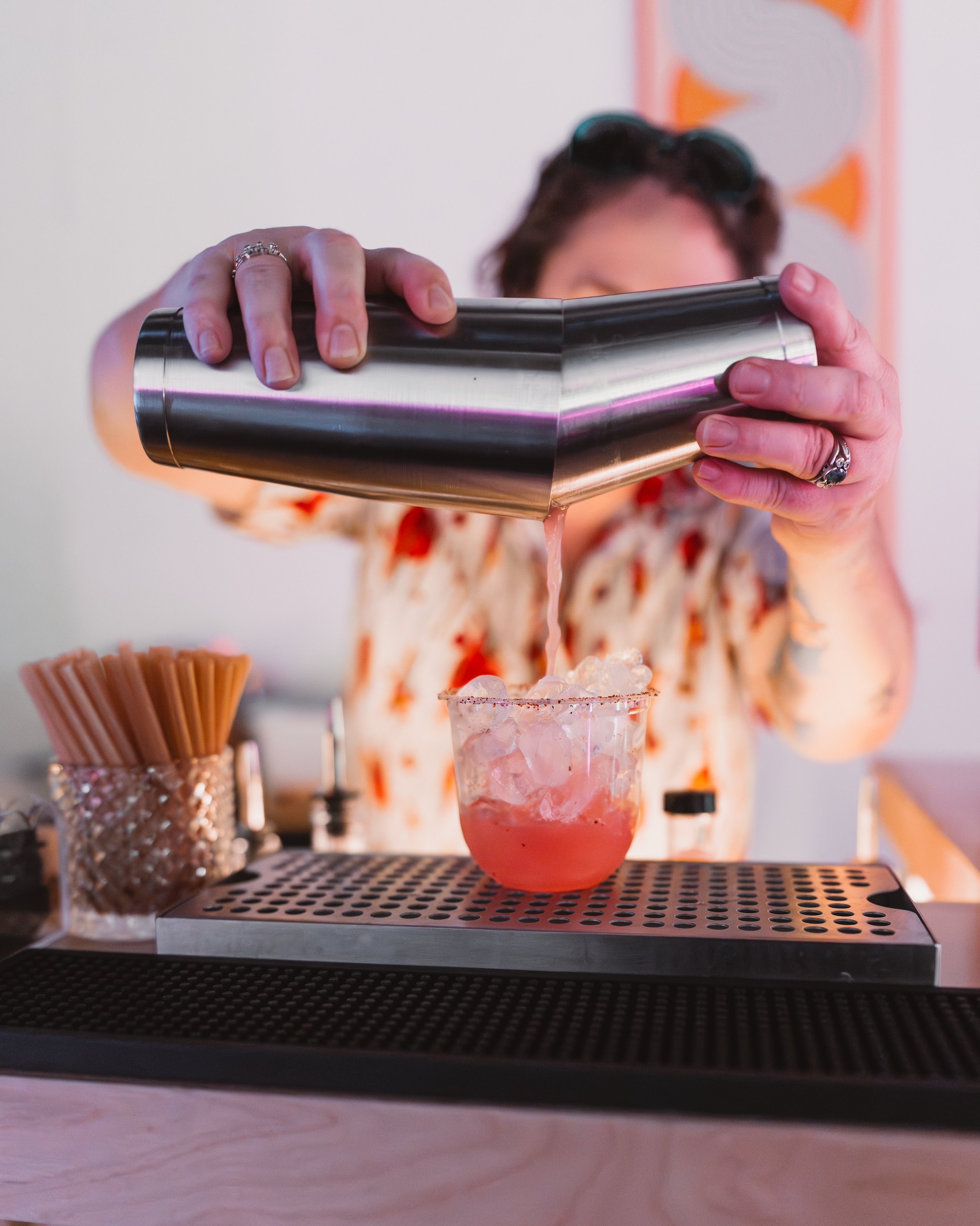 Person pouring a pink cocktail with ice into a glass at a bar, with cocktail supplies on the counter.