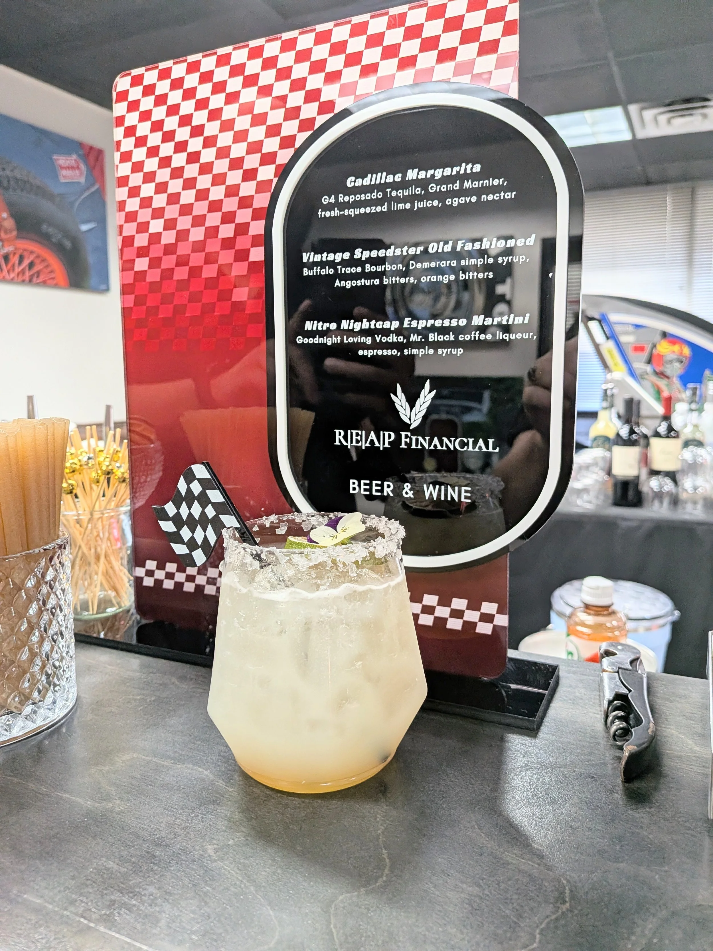 A cocktail drink with a salted rim, garnished with a flower and a small checkered flag, on a bar counter in front of a menu board listing drinks.