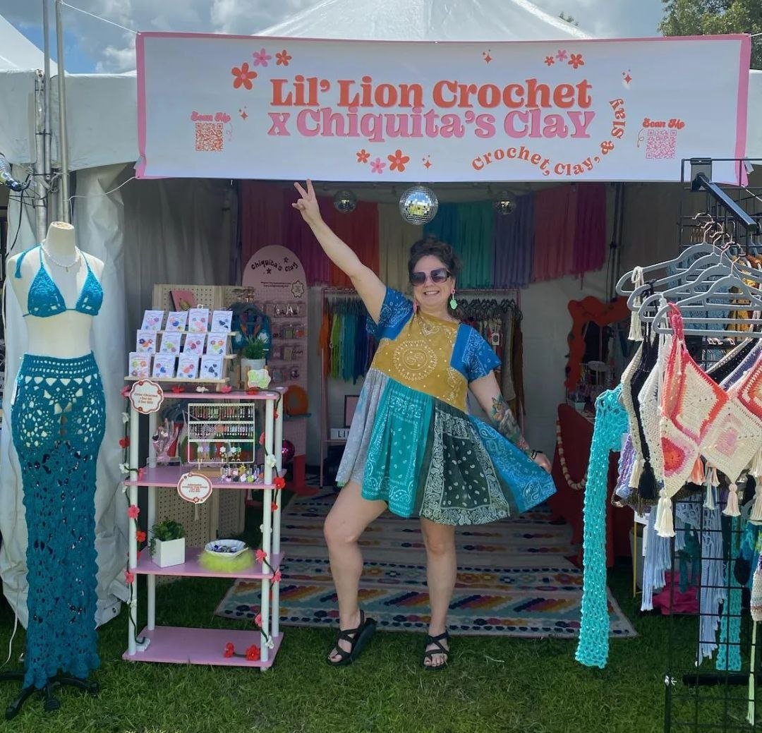 A woman standing in front of a craft booth titled 'Lil' Lion Crochet & Chiquita's Clay' at an outdoor event. She is smiling, wearing sunglasses, a colorful dress, and black sandals, and is posing with one arm raised.