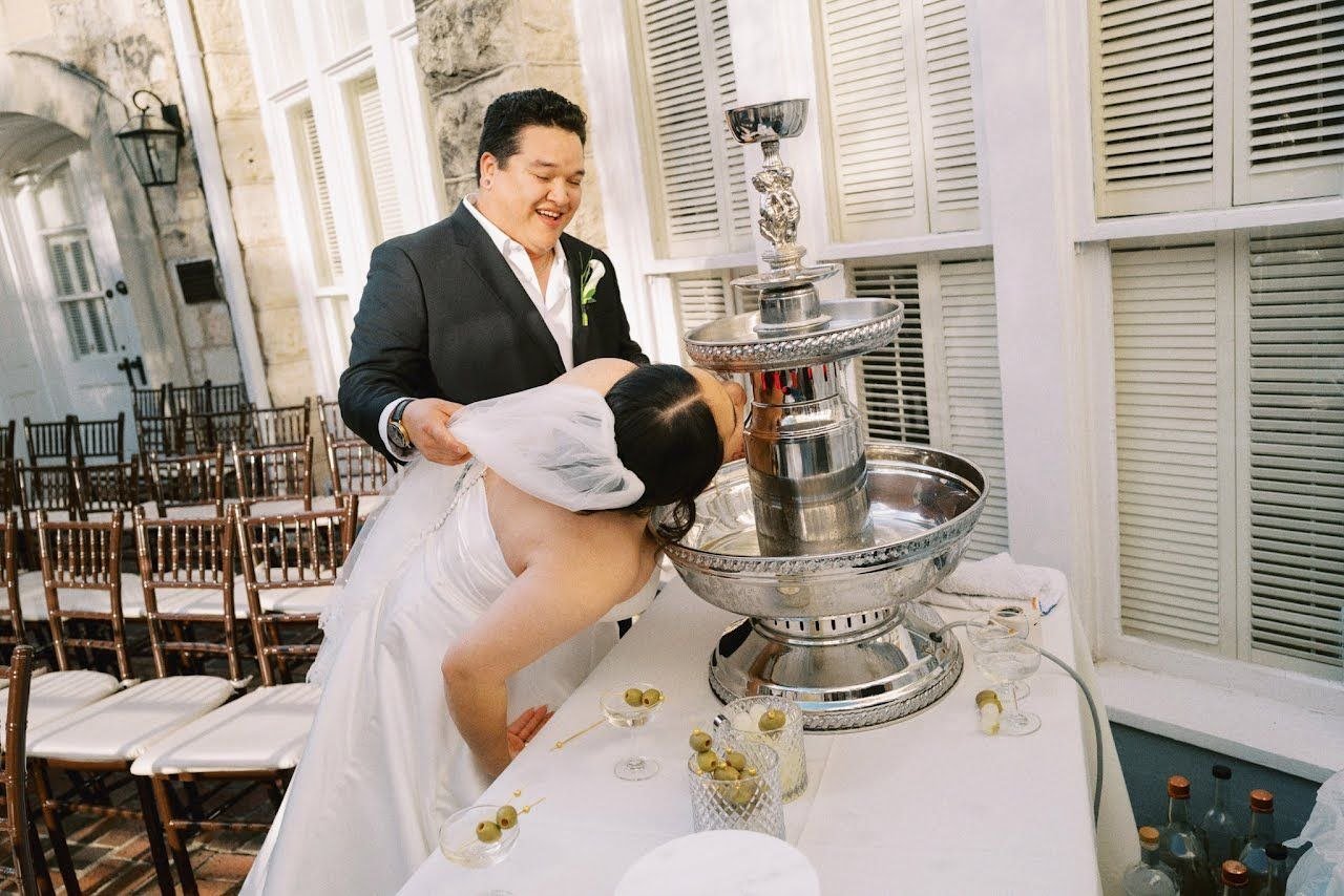 A newlywed couple participating in a wedding tradition by sipping champagne from a tiered fountain. The groom, dressed in a black suit, holds the bride, who is in a white wedding gown, as she leans over the fountain to drink.