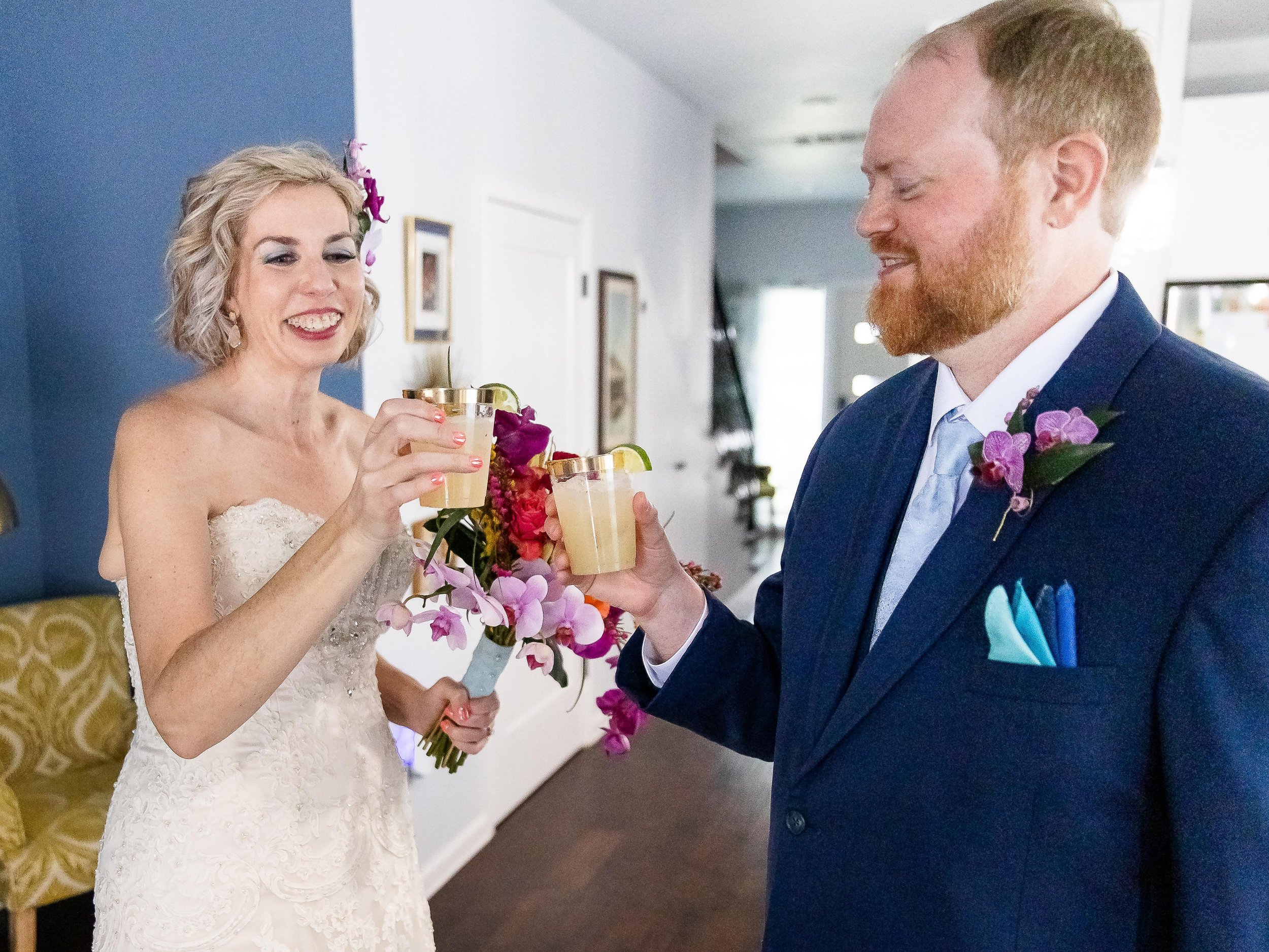 A woman in a wedding dress and a man in a suit are holding up glasses of drinks and smiling at each other. The woman has short blonde hair and is smiling with her eyes closed. The man has red hair and a beard and is smiling with his eyes closed. They are celebrating, possibly at a wedding reception, with flowers and decorations in the background.
