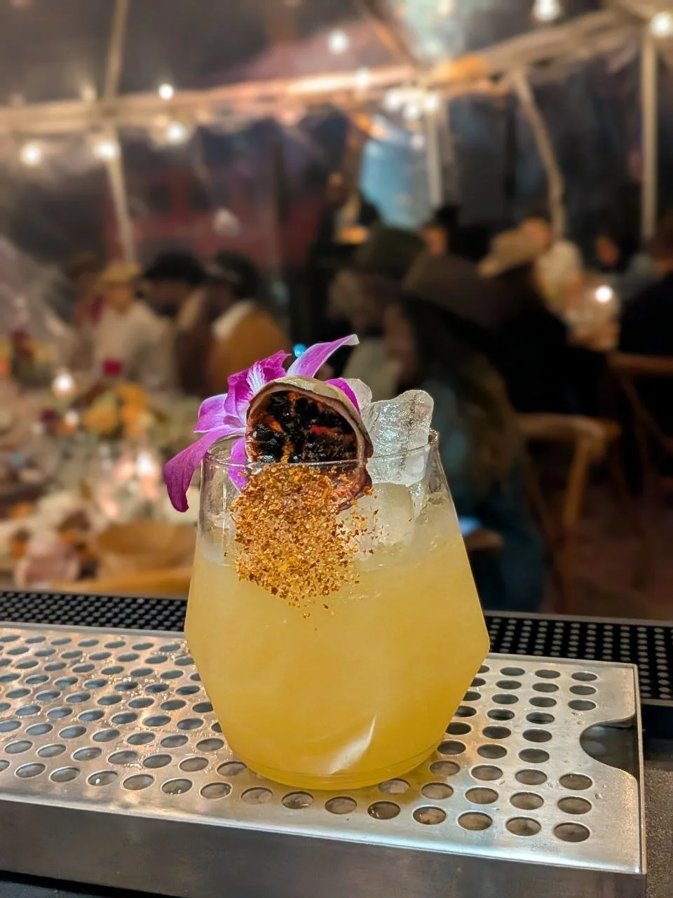 A yellow cocktail garnished with a purple orchid flower and a dried fruit slice, served with ice in a rounded glass on a metal table at a lively, dimly lit outdoor setting.