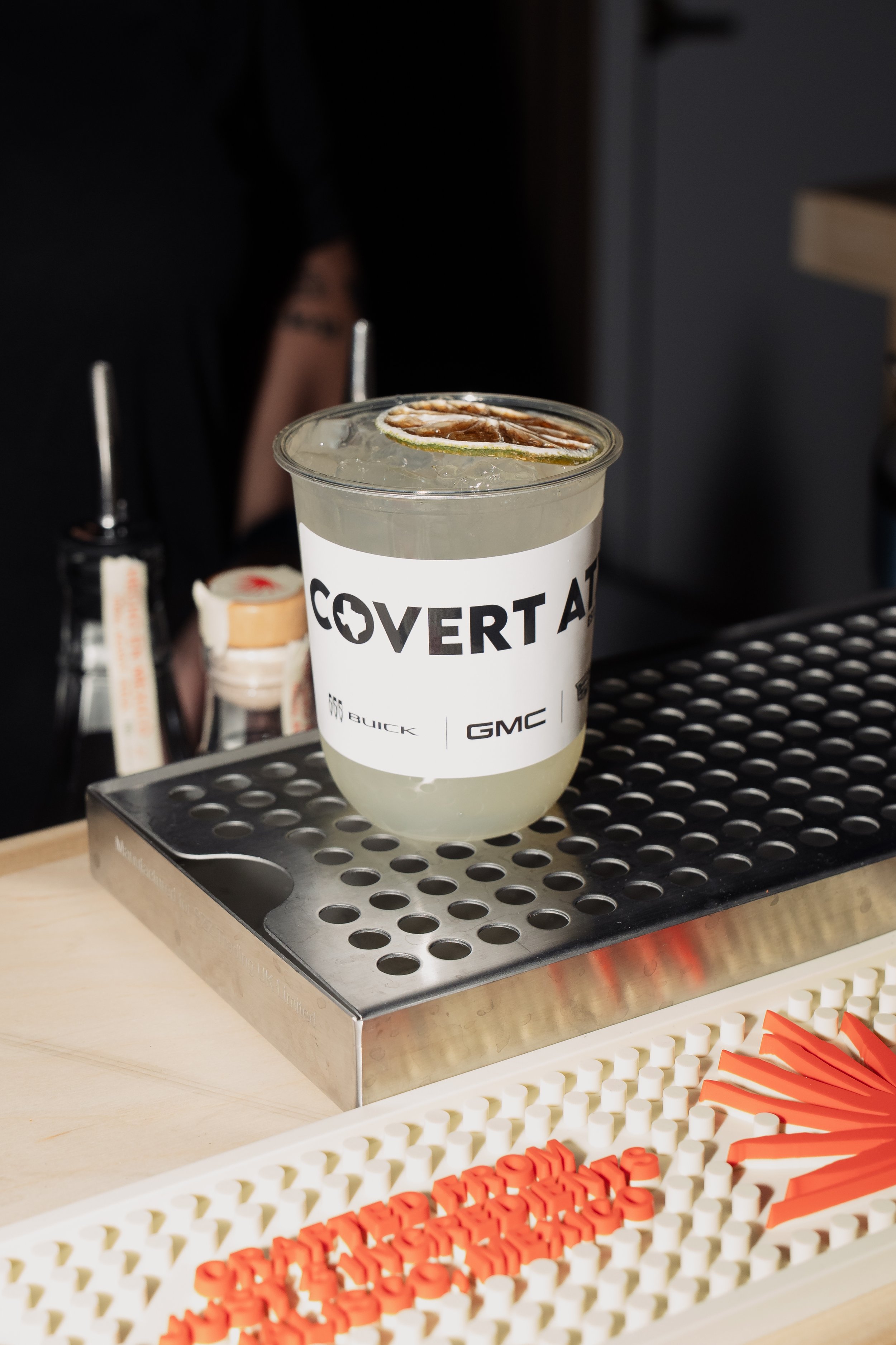 A clear plastic cup with a lemon-lime soda drink garnished with a lime and lemon slice, placed on a metal bar mat.
