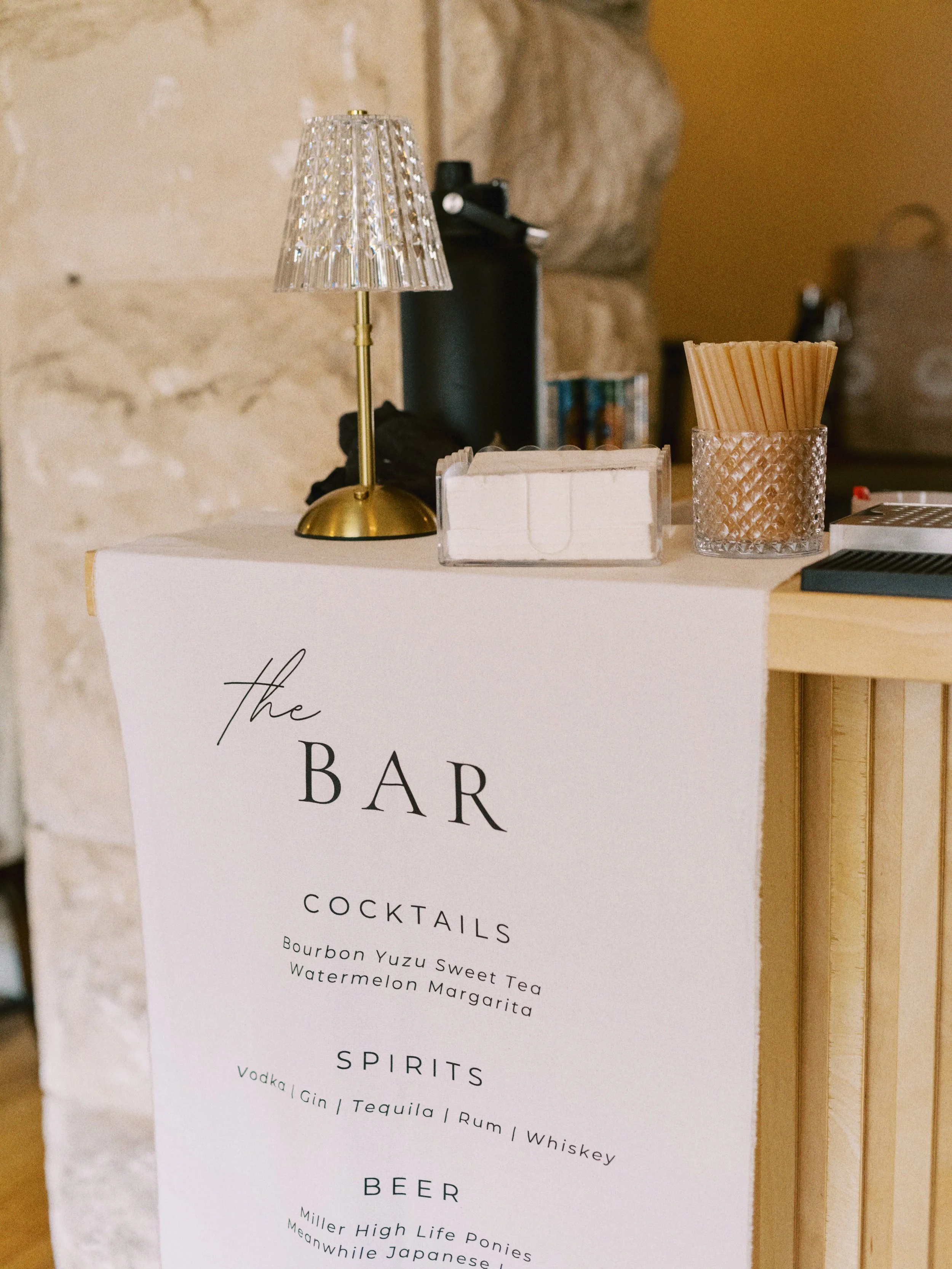 A menu on a table lists drinks including cocktails, spirits, and beer, with a small crystal lamp, a glass container with straws, and other items on the table.
