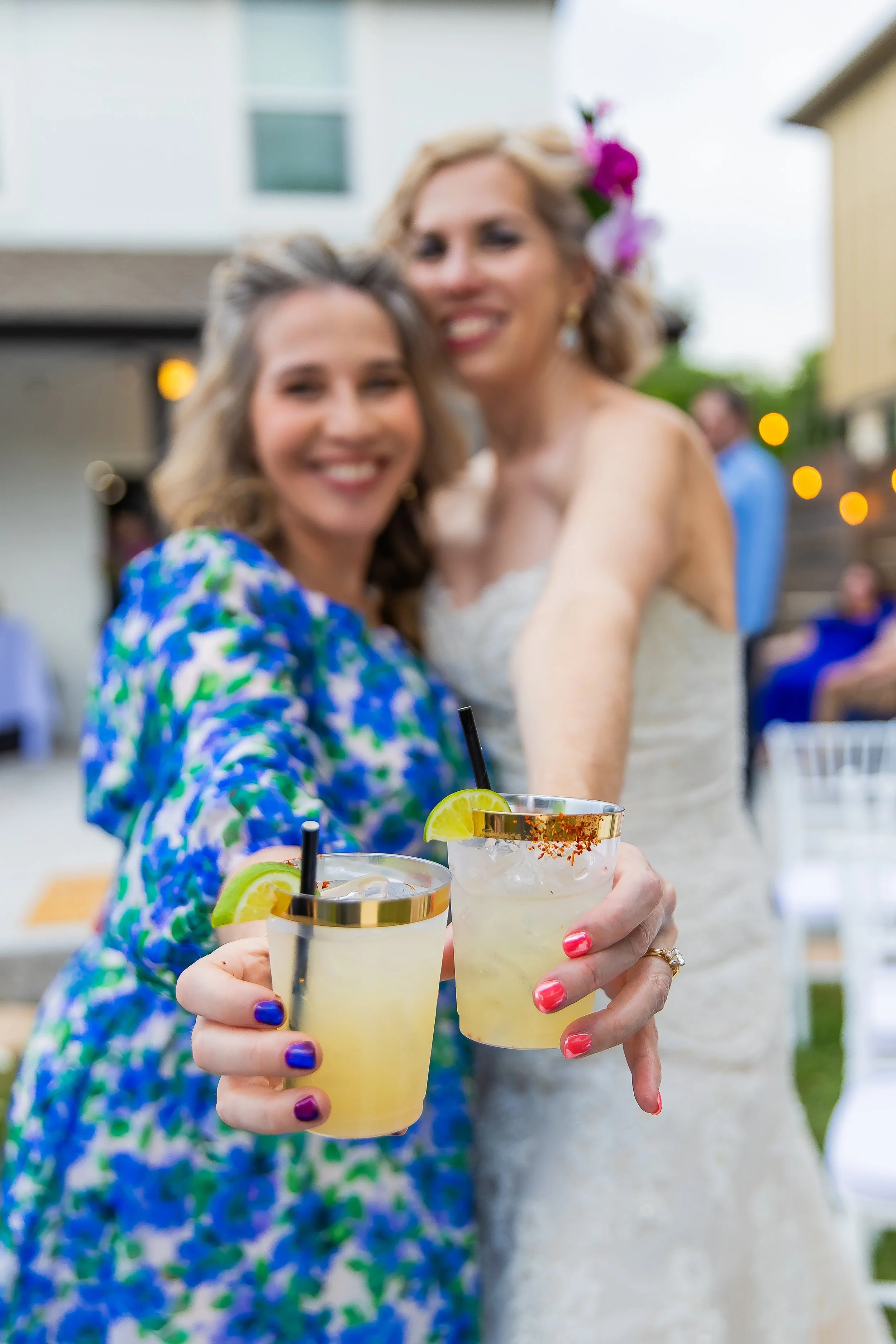 Two women smiling and holding cocktails with lemon slices, salt rim, and colorful nails, at an outdoor gathering during dusk.