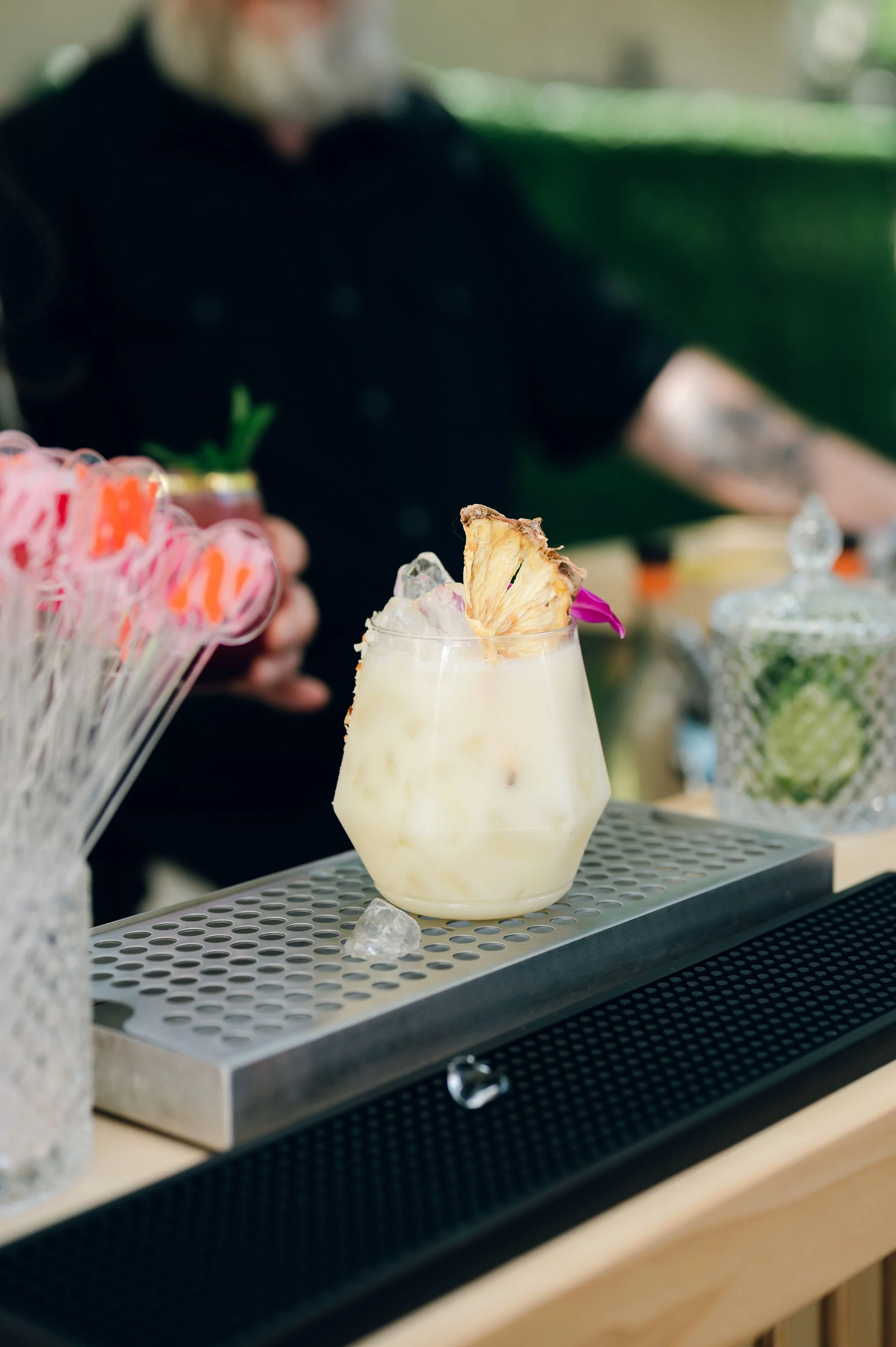 A tropical cocktail garnished with a pineapple slice and pink flower, placed on a bar counter with ice cubes and other decorations.