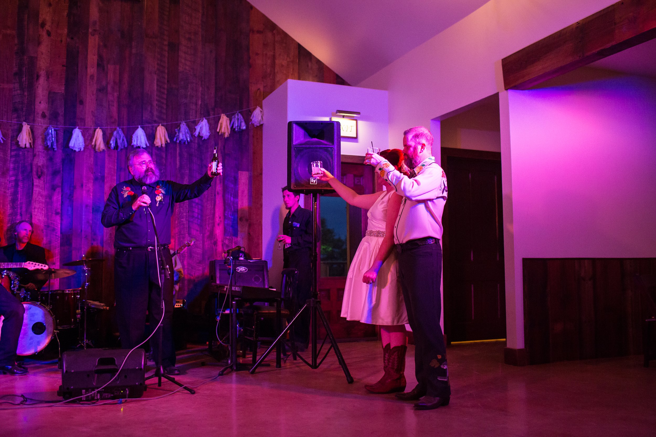 People celebrating at a wedding reception. A man with a beard and glasses holds a beer and a microphone while standing on a stage. An Austin bride and groom in a light-colored dress and cowboy boots, and a man in a white shirt and black pants, are to
