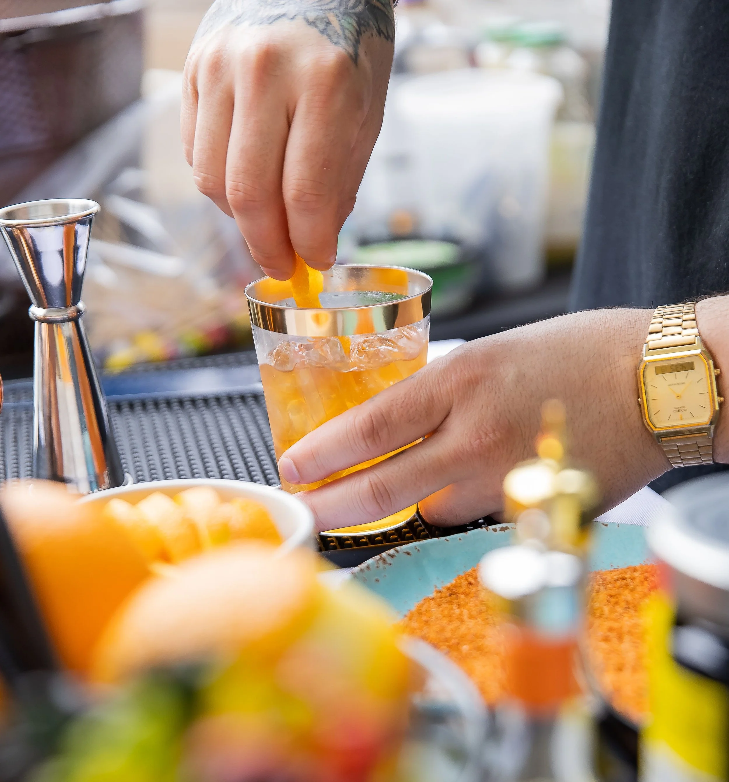 A person is placing a lemon twist into a cocktail glass filled with a yellow-colored drink, with a wristwatch visible on their left wrist.