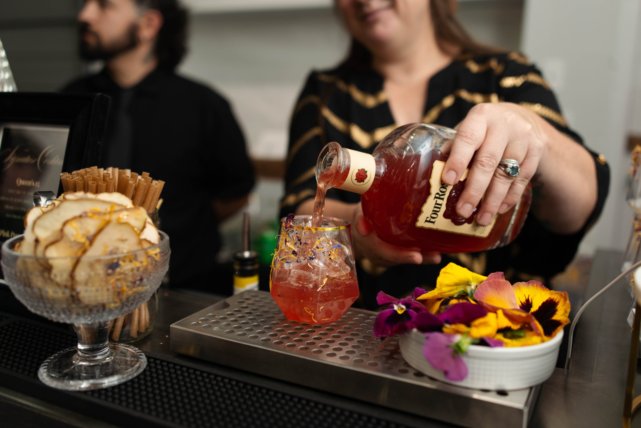A woman pouring a signature pink cocktail from a bottle into a glass with ice, with a man in the background at a bar or restaurant.