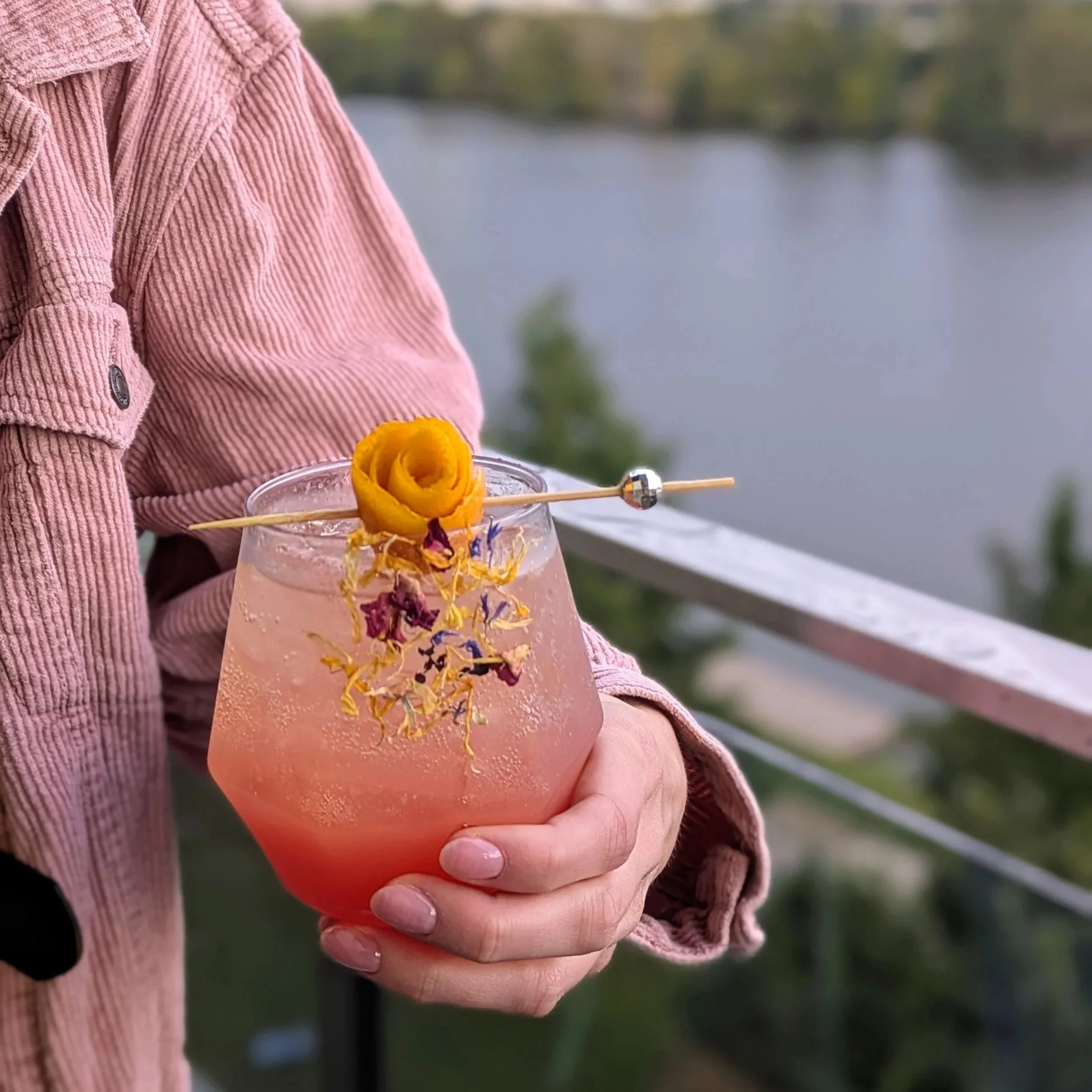 Social Butterfly ATX signature cocktail garnished with a citrus rose, edible flowers, and a cocktail pick, held overlooking Lady Bird Lake and the Austin skyline