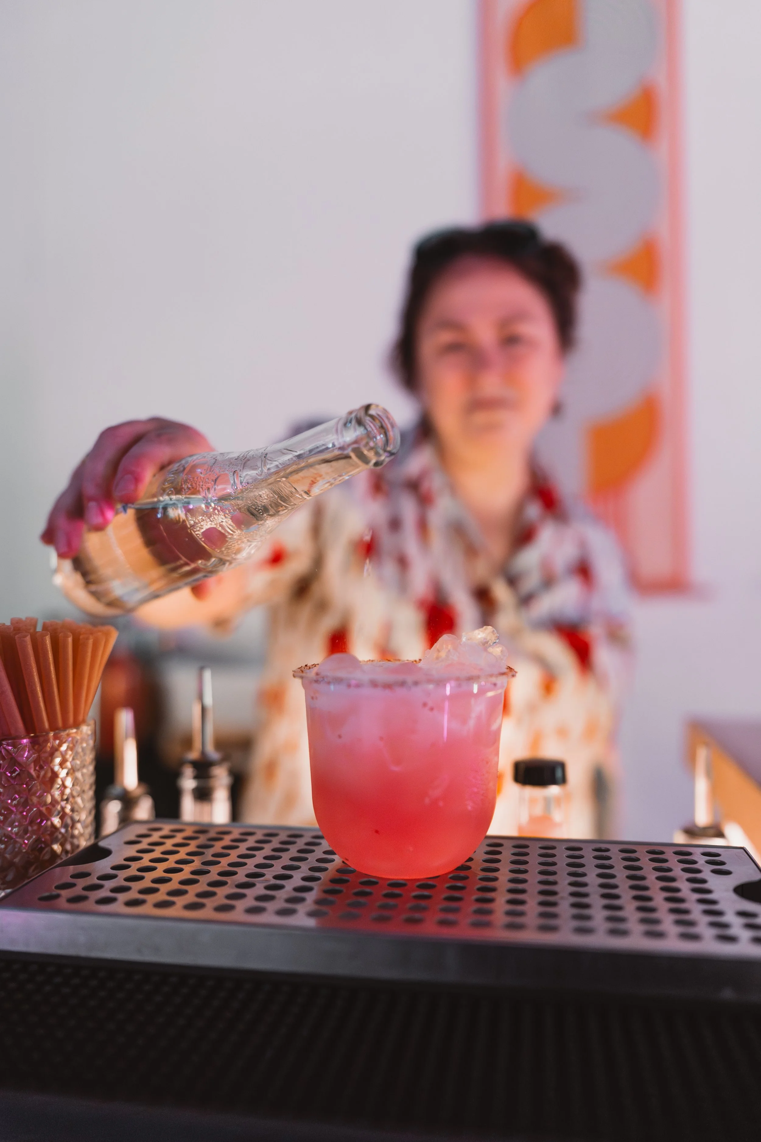 A woman in a floral shirt pours liquor into a pink cocktail.