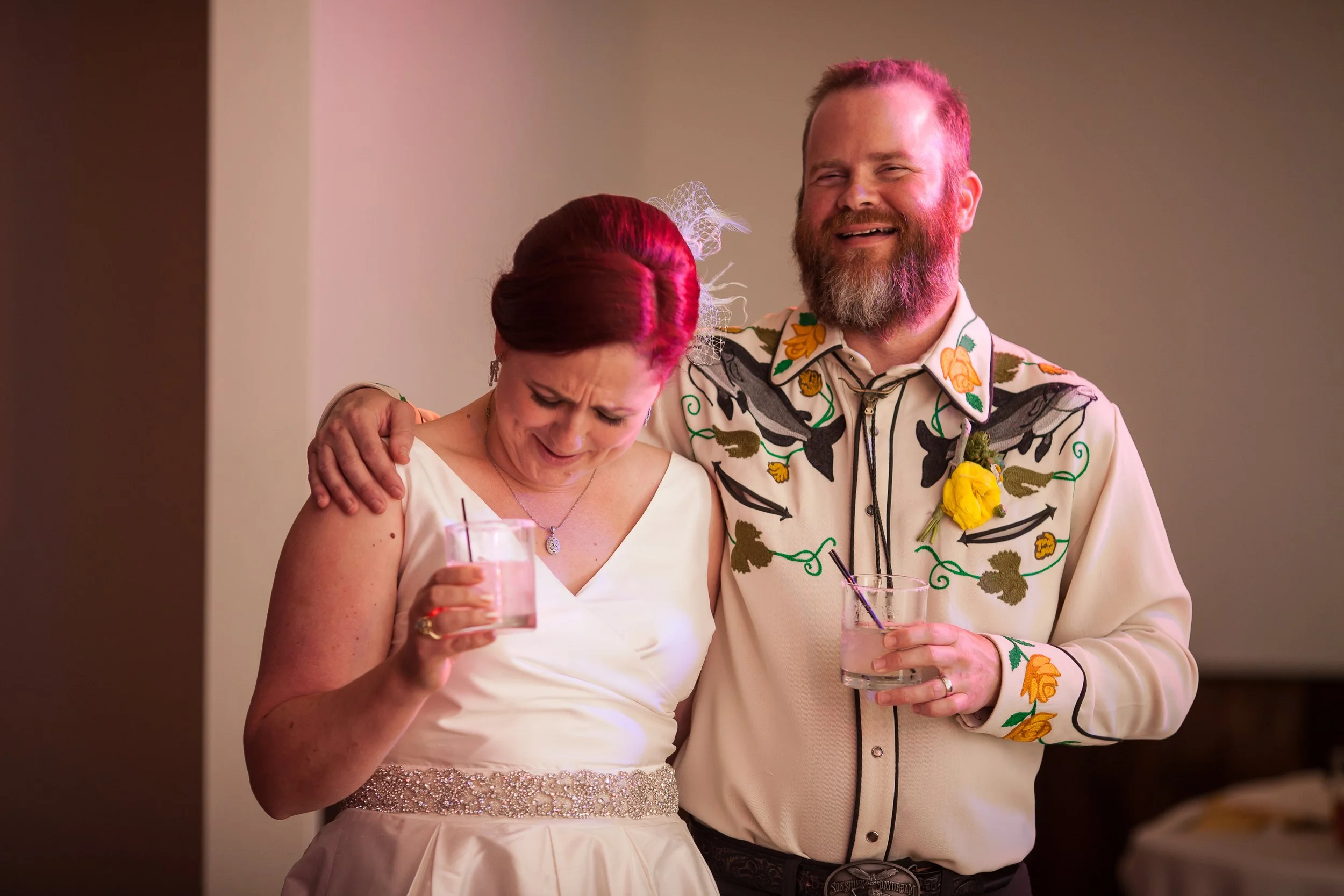 A man with a beard and a woman with red hair at a mid-sized wedding celebration. The woman appears emotional, holding a glass, and the man has his arm around her shoulder, smiling and holding a drink. They are dressed in semi-formal attire, and the w