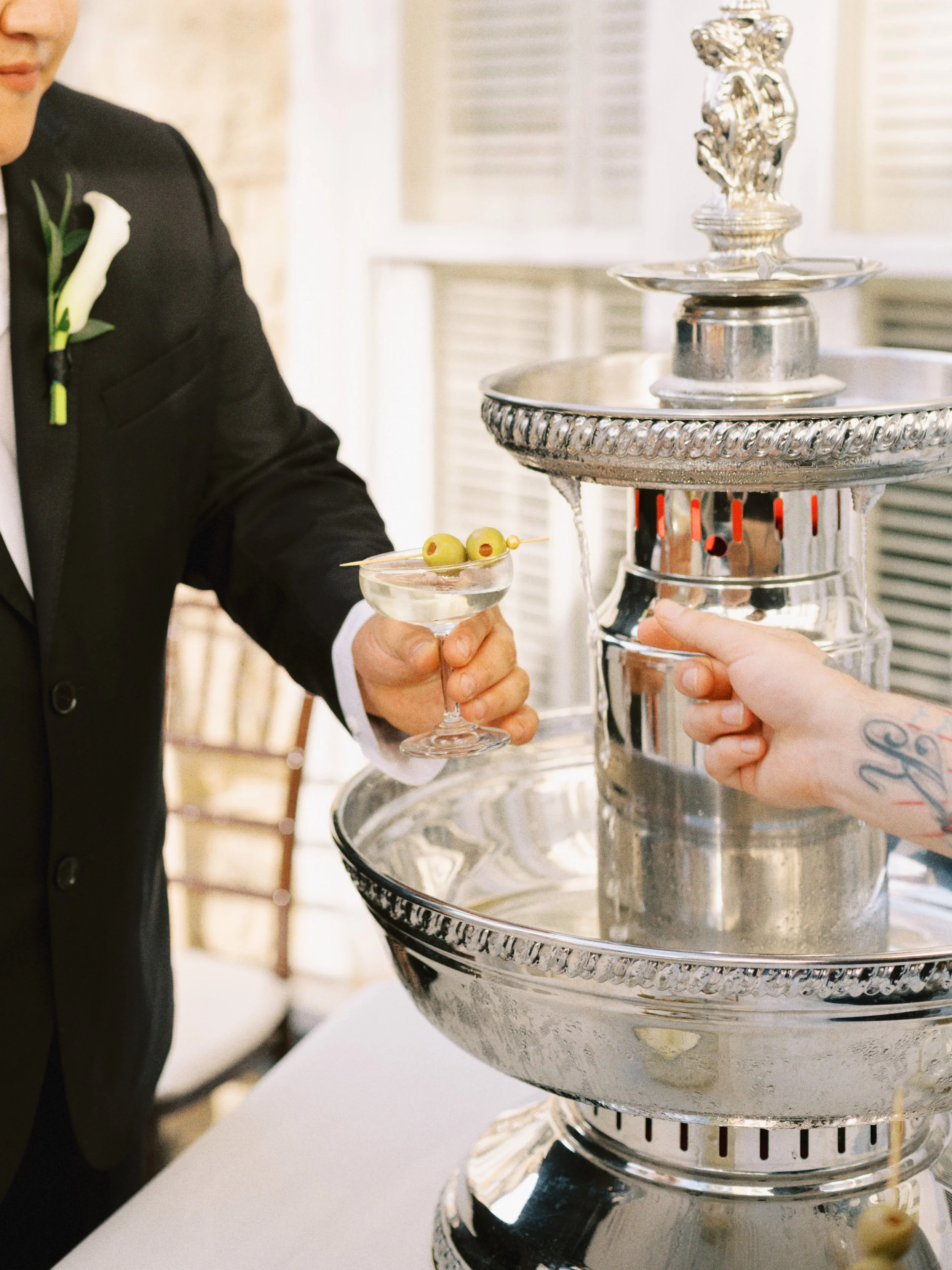 A person in formal attire is holding a martini glass with green olives, standing next to a silver chafing dish with a decorative sculpture on top, in an indoor setting.