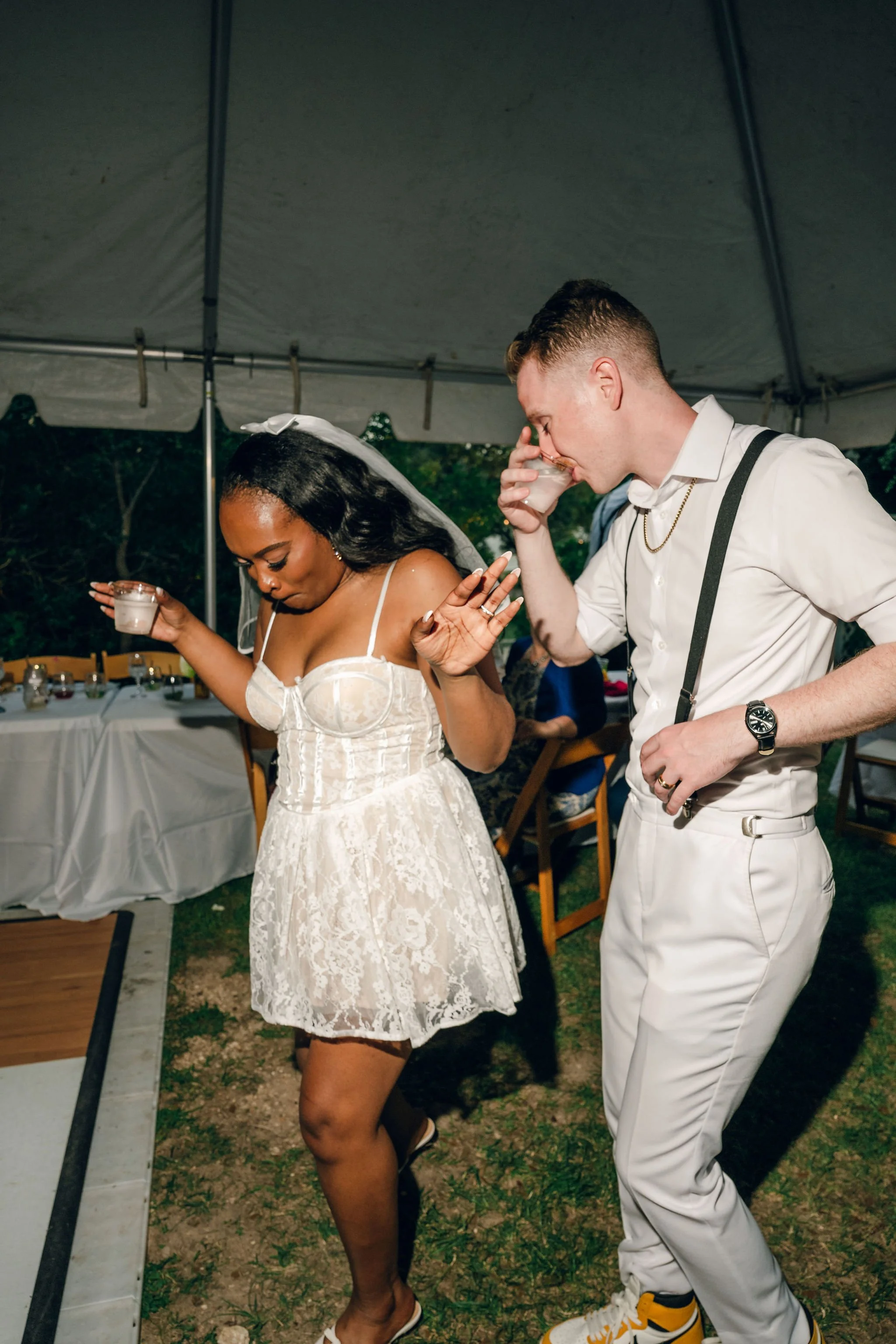A woman in a white lace dress and a man in a white shirt and pants are eating at a gathering under a tent. The woman has a veil and the man has suspenders. They are drinking from small cups, possibly at a wedding celebration.