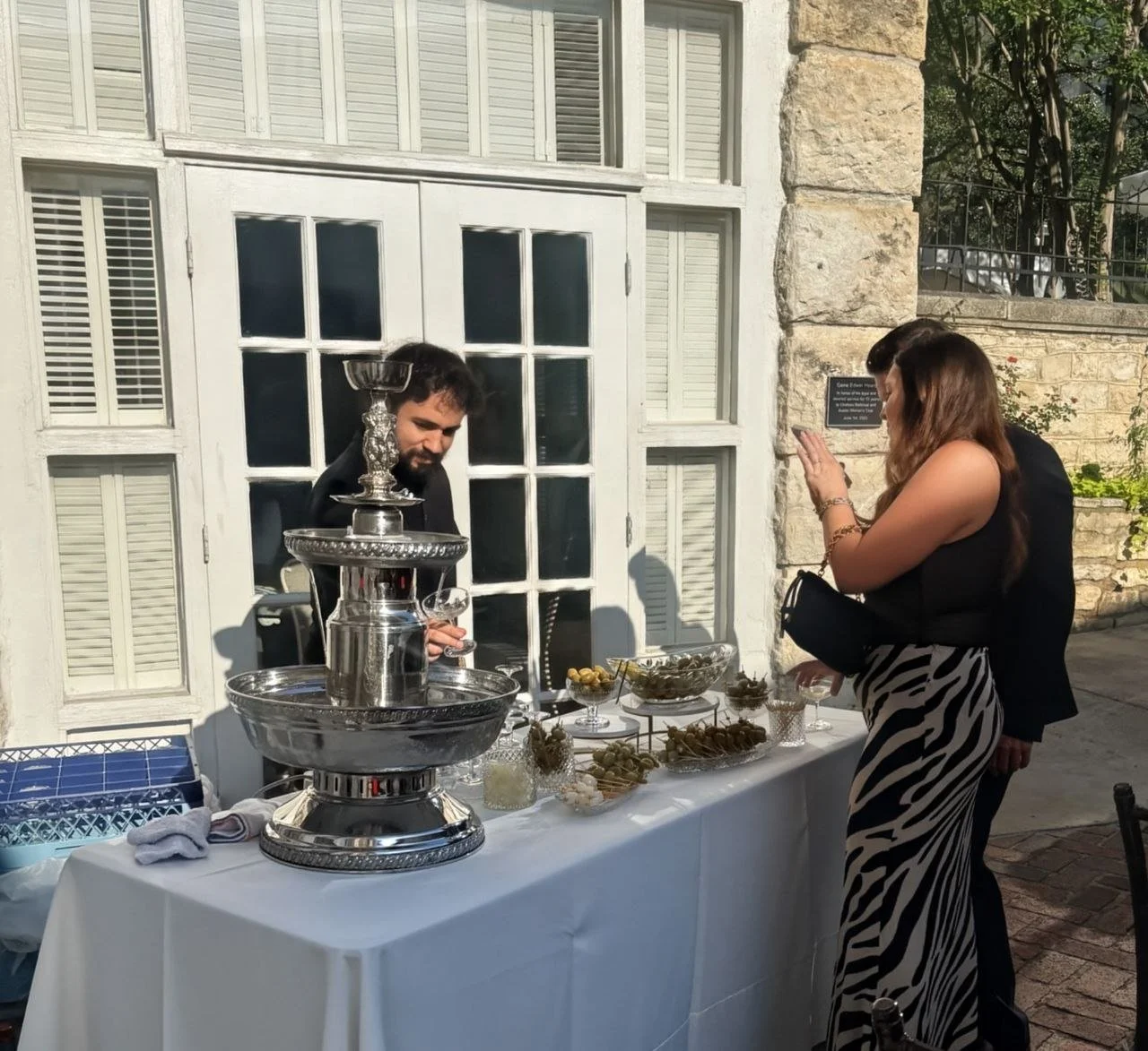 People at an outdoor event with a table of food and a silver water fountain, standing near a stone wall and white window pane door.