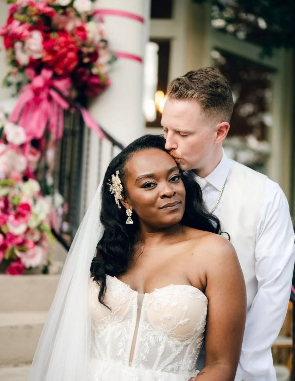 A newlywed couple on their wedding day. The groom is kissing the bride's forehead while she faces the camera with a serious expression. The bride is wearing a strapless lace wedding dress and earrings, and the groom is dressed in a white shirt with a