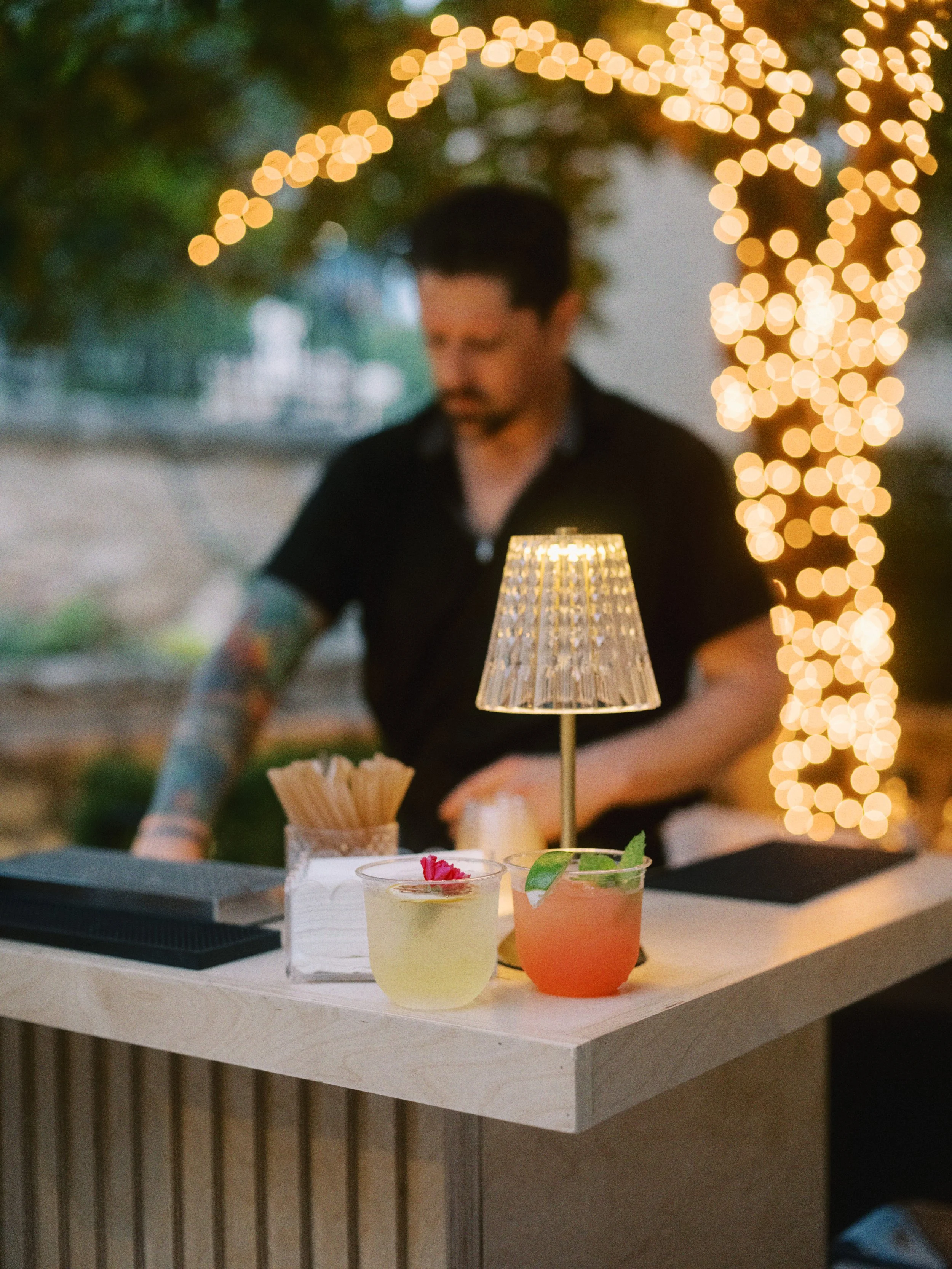 A person standing behind a table with two cocktails, candles, and decorations, with bokeh lights in the background.