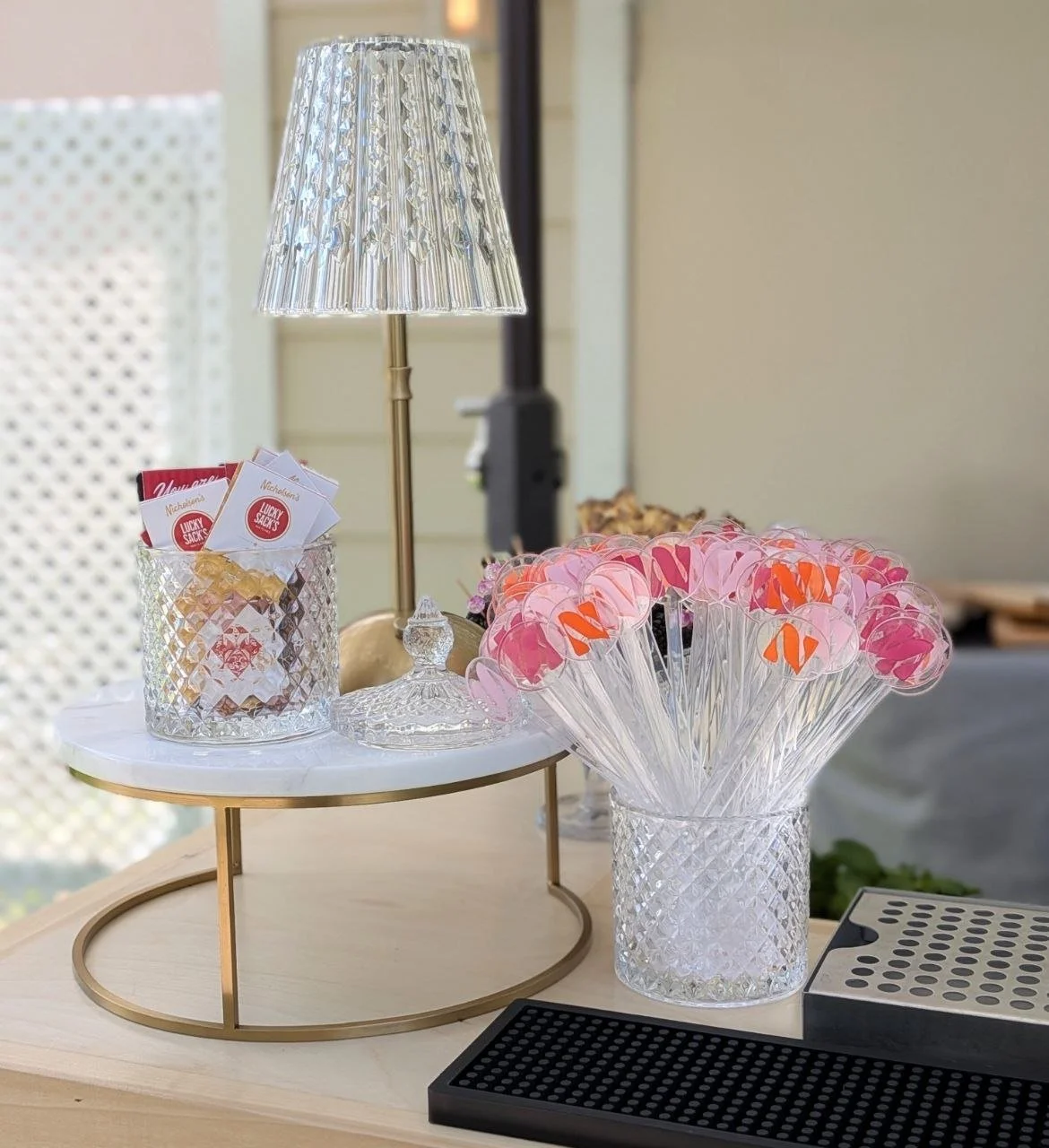 Decorative table display with pink and orange-themed items, including lollipops in a glass vase, a two-tiered tray with small packets, and a crystal lamp with a silver base.